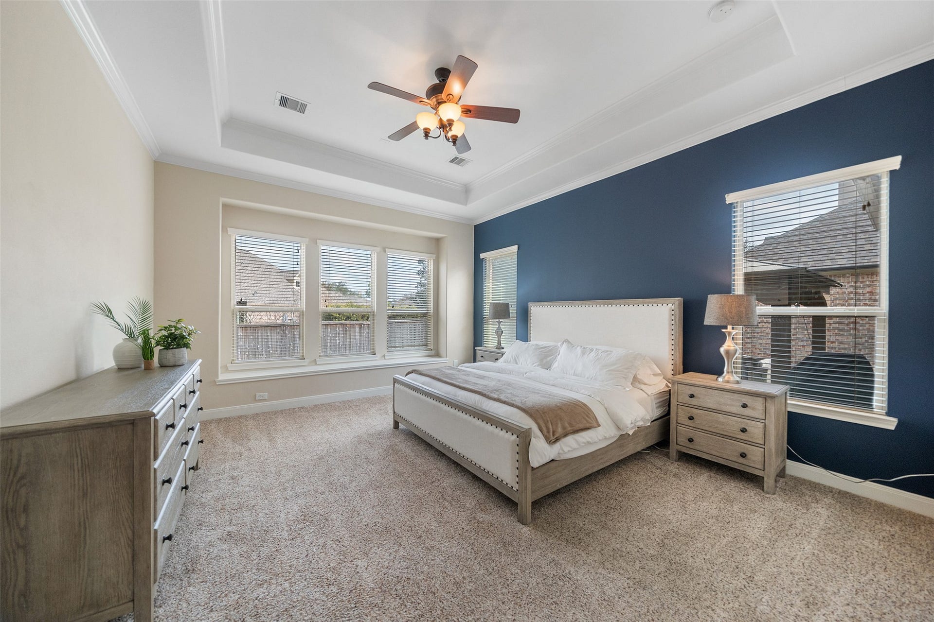 34 Danby Place, The Woodlands, TX 77375 Spacious primary bedroom with a cozy carpet, featuring a large bed, elegant furniture, and a ceiling fan. Natural light streams through multiple windows, complementing the soothing blue accent wall. It includes a neutral color palette with a tray ceiling and a dresser for storage. Perfect for comfortable living.