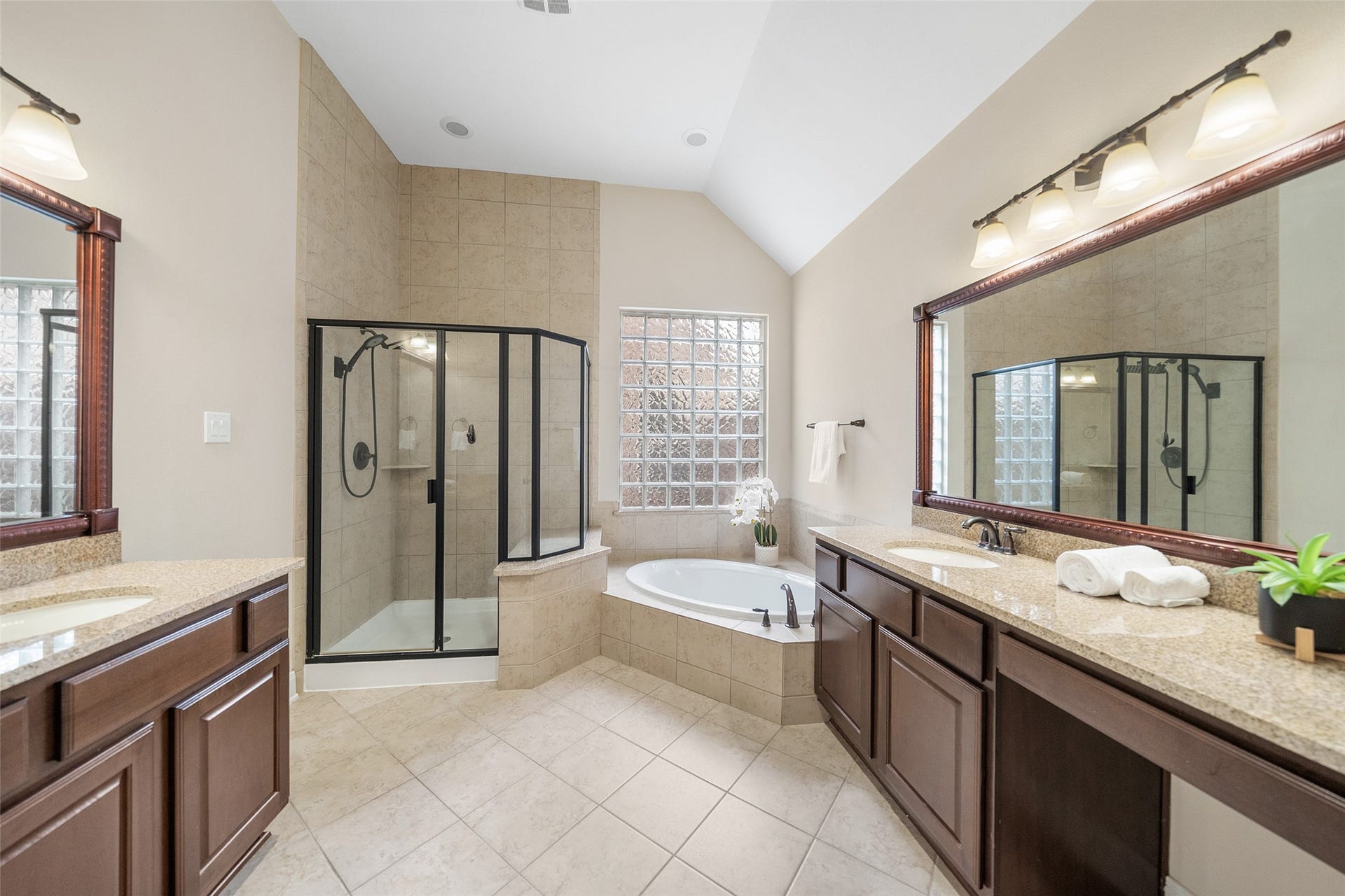 34 Danby Place, The Woodlands, TX 77375 This spacious primary bathroom features a large corner bathtub, a separate glass-enclosed shower, dual sinks with granite countertops, and ample cabinetry. Natural light filters through glass block windows, enhancing the elegant tile flooring and soft neutral tones, creating a bright and modern retreat.