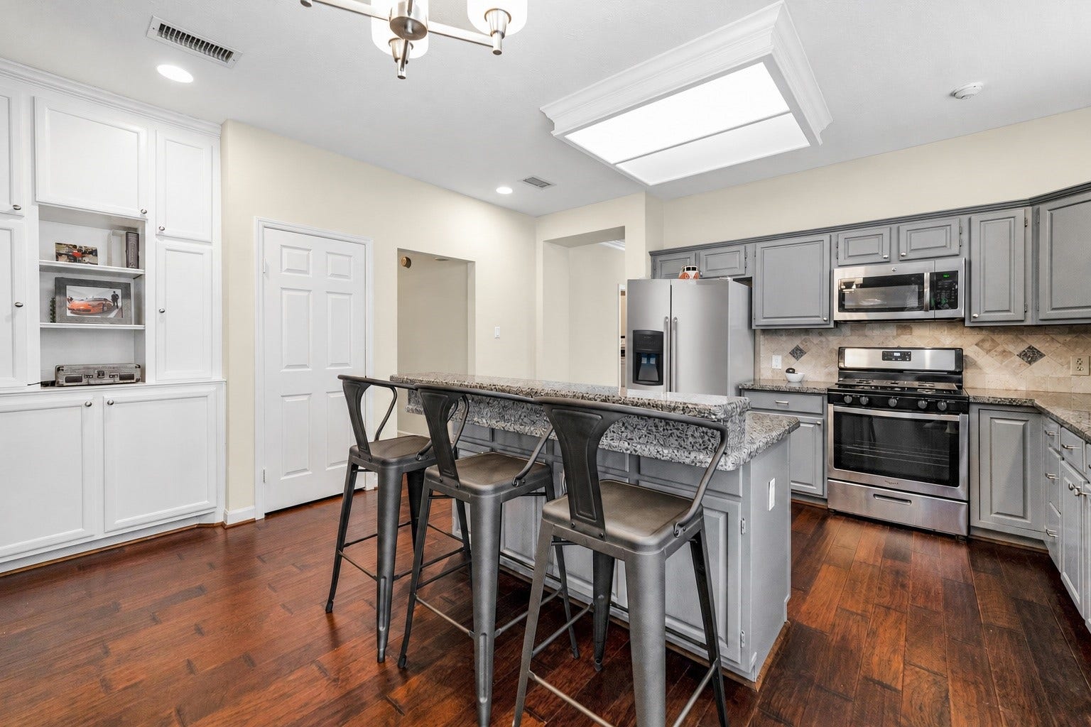 11 Sweetdream Place, The Woodlands, TX 77381 Charming kitchen with modern gray cabinetry, stainless steel appliances, and a spacious granite-topped island that offers casual seating. Warm wood flooring enhances the inviting atmosphere, complemented by built-in shelving for additional storage.