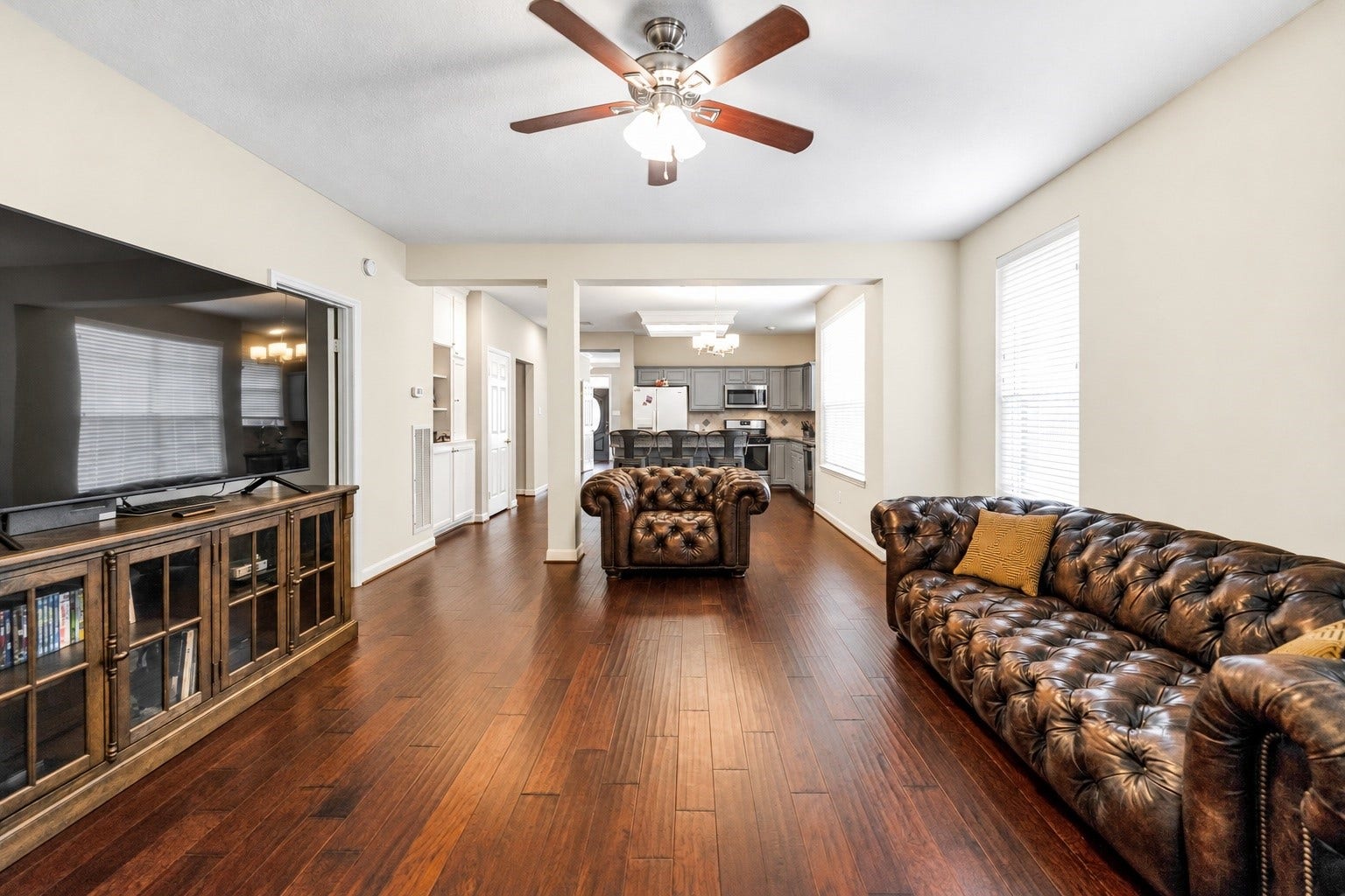 11 Sweetdream Place, The Woodlands, TX 77381 Spacious living area with rich hardwood floors, elegant leather seating, and a ceiling fan, seamlessly flowing into a modern kitchen with stainless steel appliances.