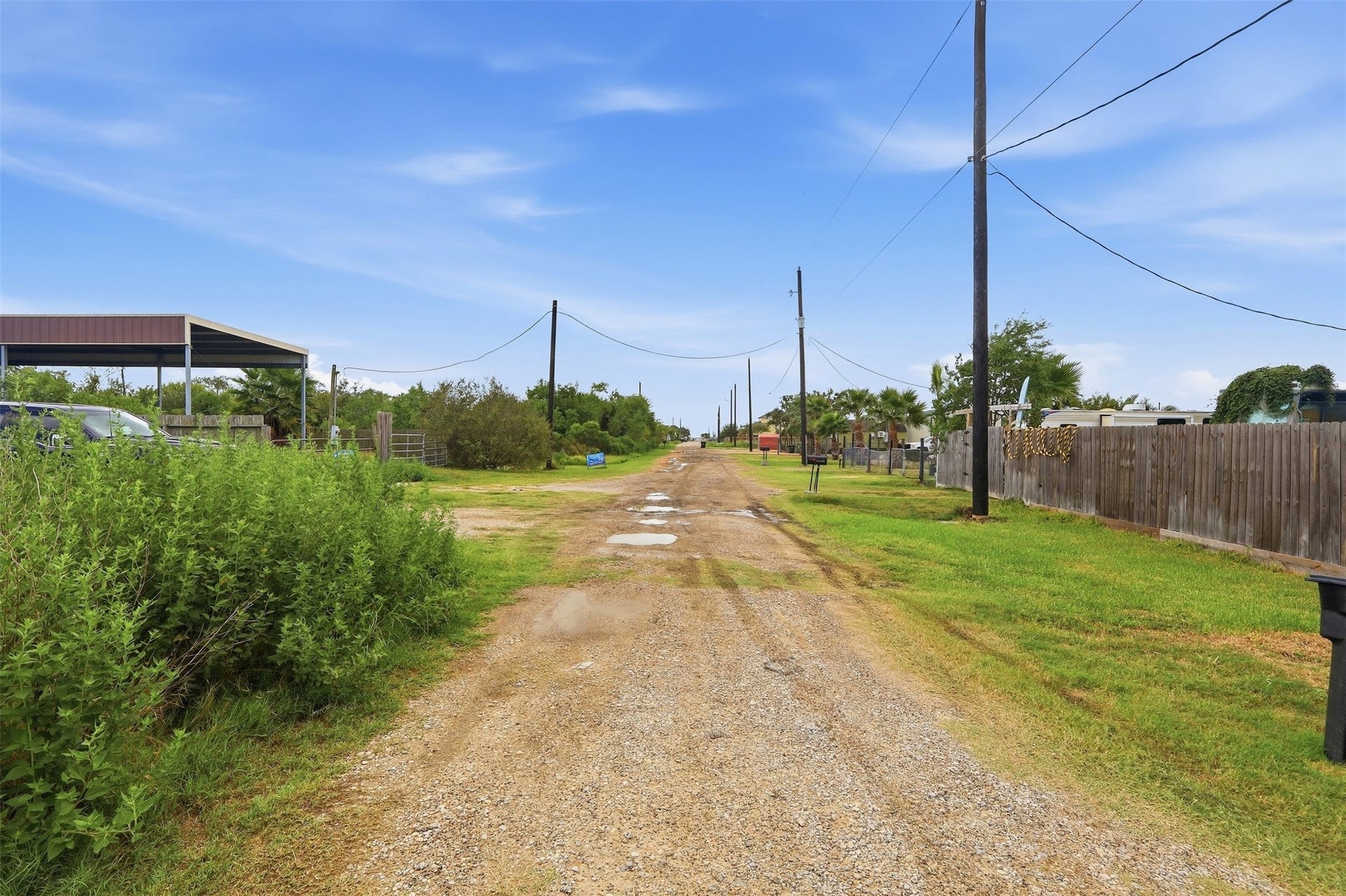 17 W Street, San Leon, TX 77539 Gravel side road running along the property, with utility poles overhead. Neighboring lots are fenced and developed, and the street leads in the direction of the bay.