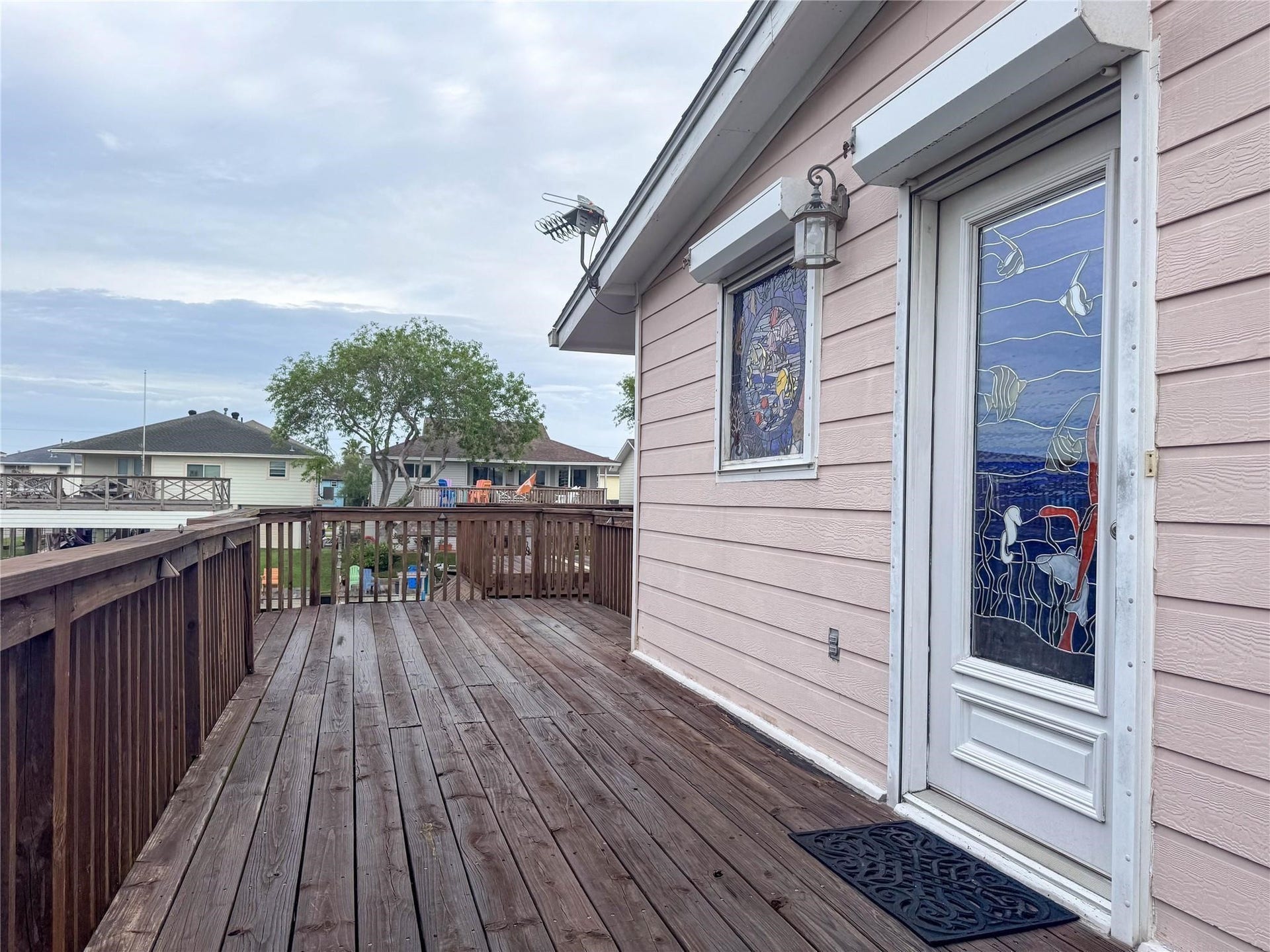 1031 Redfish Street, Bayou Vista, TX 77563 This photo showcases a charming deck with wooden flooring, adjacent to a house with light-colored siding. It features a decorative glass door and window, offering a blend of privacy and artistic flair. The view includes neighboring homes and greenery, indicating a cozy neighborhood setting.