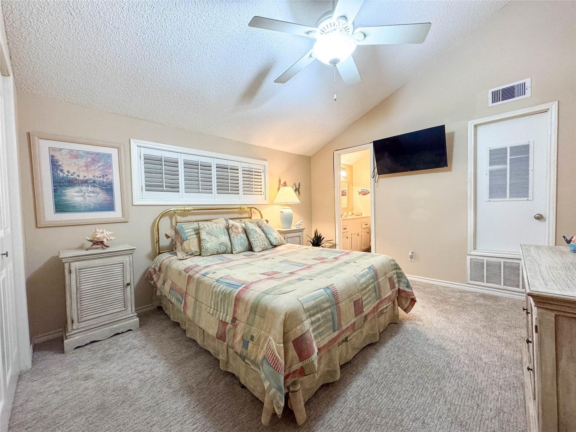 1031 Redfish Street, Bayou Vista, TX 77563 Cozy bedroom with soft lighting, featuring a comfortable bed, ceiling fan, and neutral decor. The room includes a wall-mounted TV, adjacent bathroom, and ample natural light from a window with shutters.