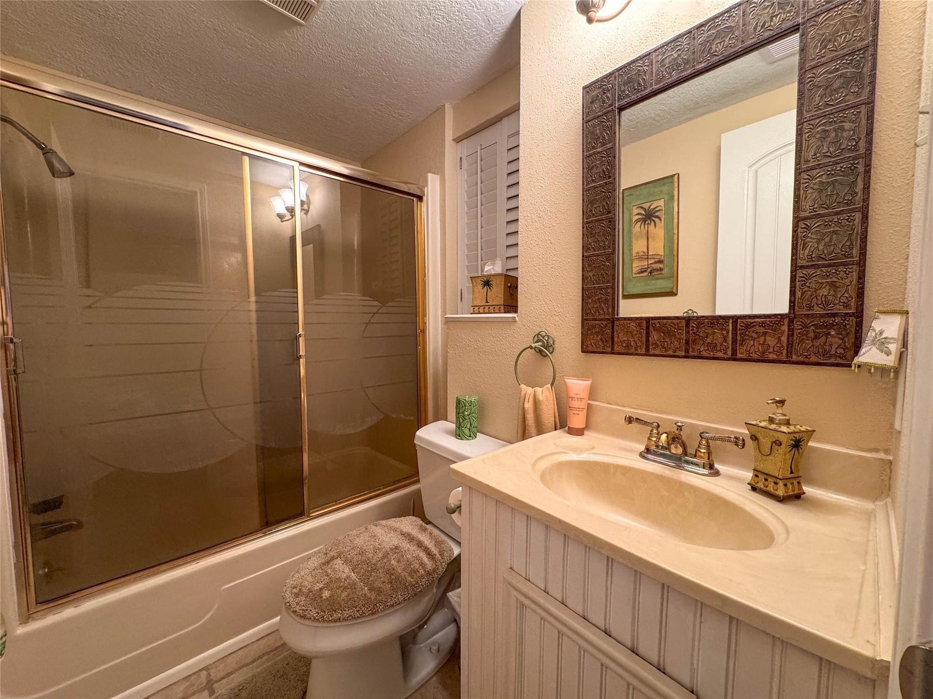 1031 Redfish Street, Bayou Vista, TX 77563 This cozy bathroom features a bathtub with sliding glass doors, a toilet with a plush cover, and a classic vanity with a decorative framed mirror. The warm lighting and palm-themed decor add a touch of elegance.