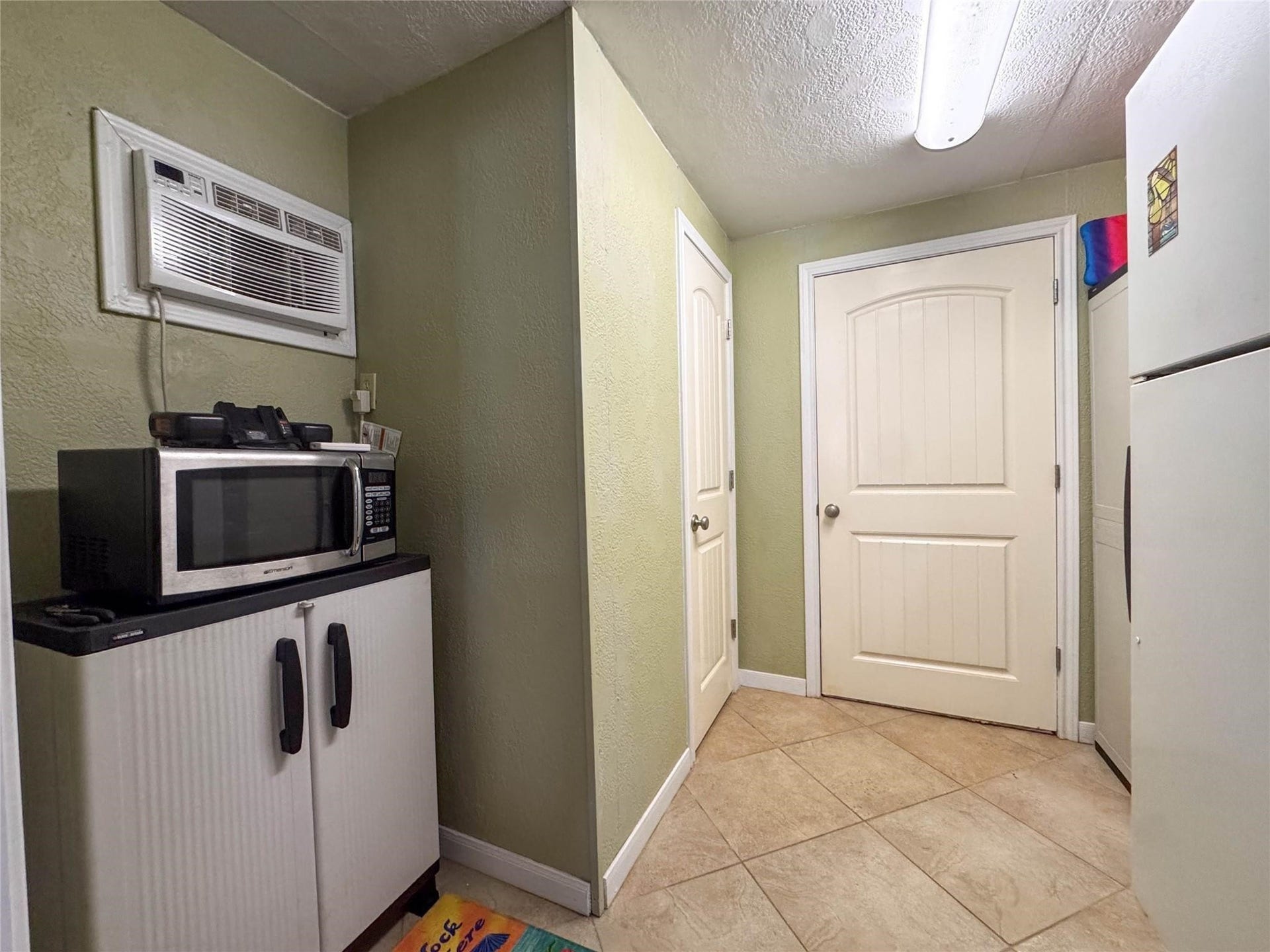 1031 Redfish Street, Bayou Vista, TX 77563 Compact utility space with tile flooring, featuring a wall-mounted air conditioner, a microwave atop a storage cabinet, and two white doors. Neutral wall color and ceiling lighting create a simple, functional area.