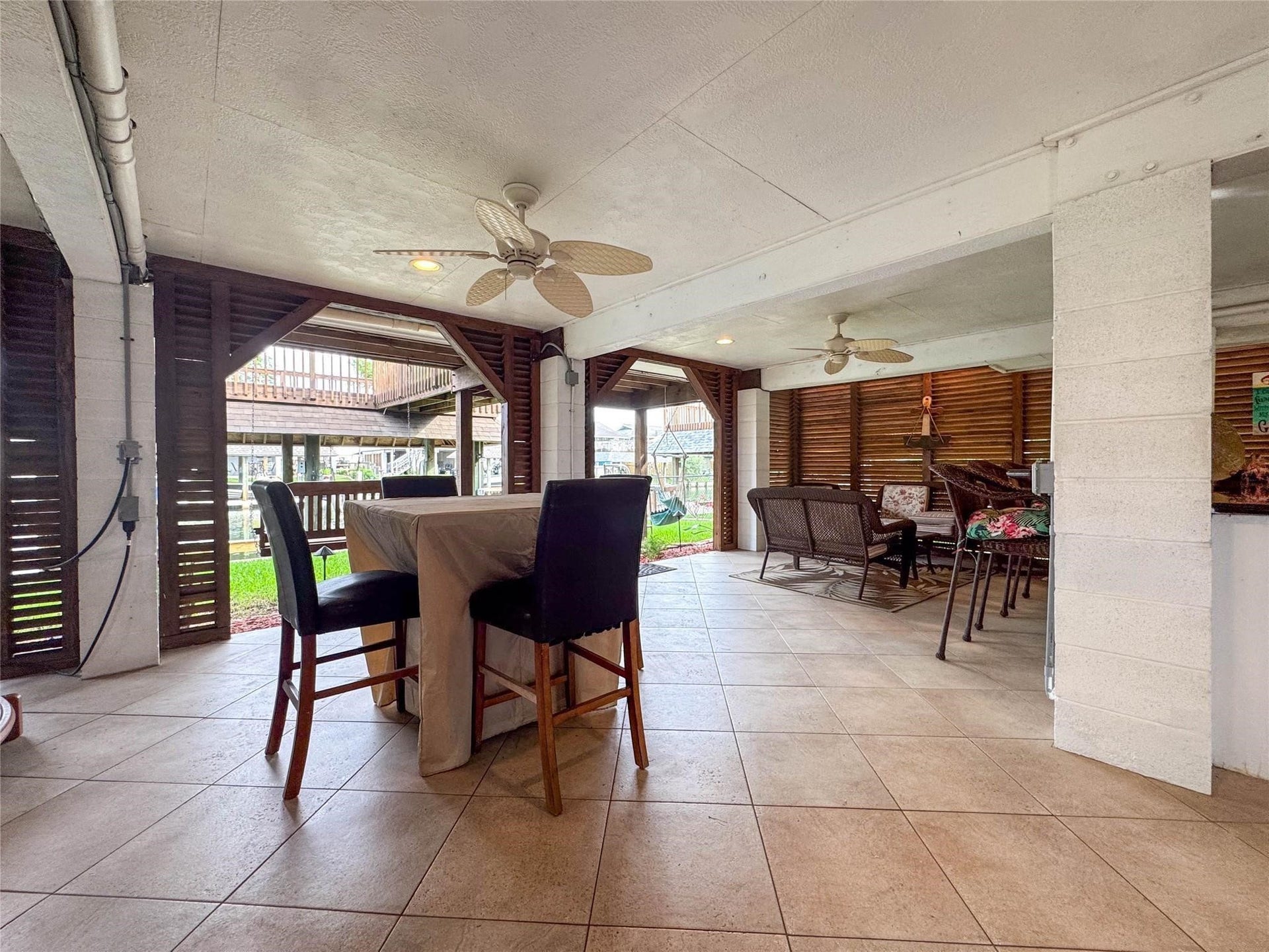 1031 Redfish Street, Bayou Vista, TX 77563 This spacious covered patio features tiled flooring, ceiling fans, and ample seating areas, perfect for outdoor entertaining. It offers a cozy, open-air feel with wooden slat walls and a view of the surrounding greenery.