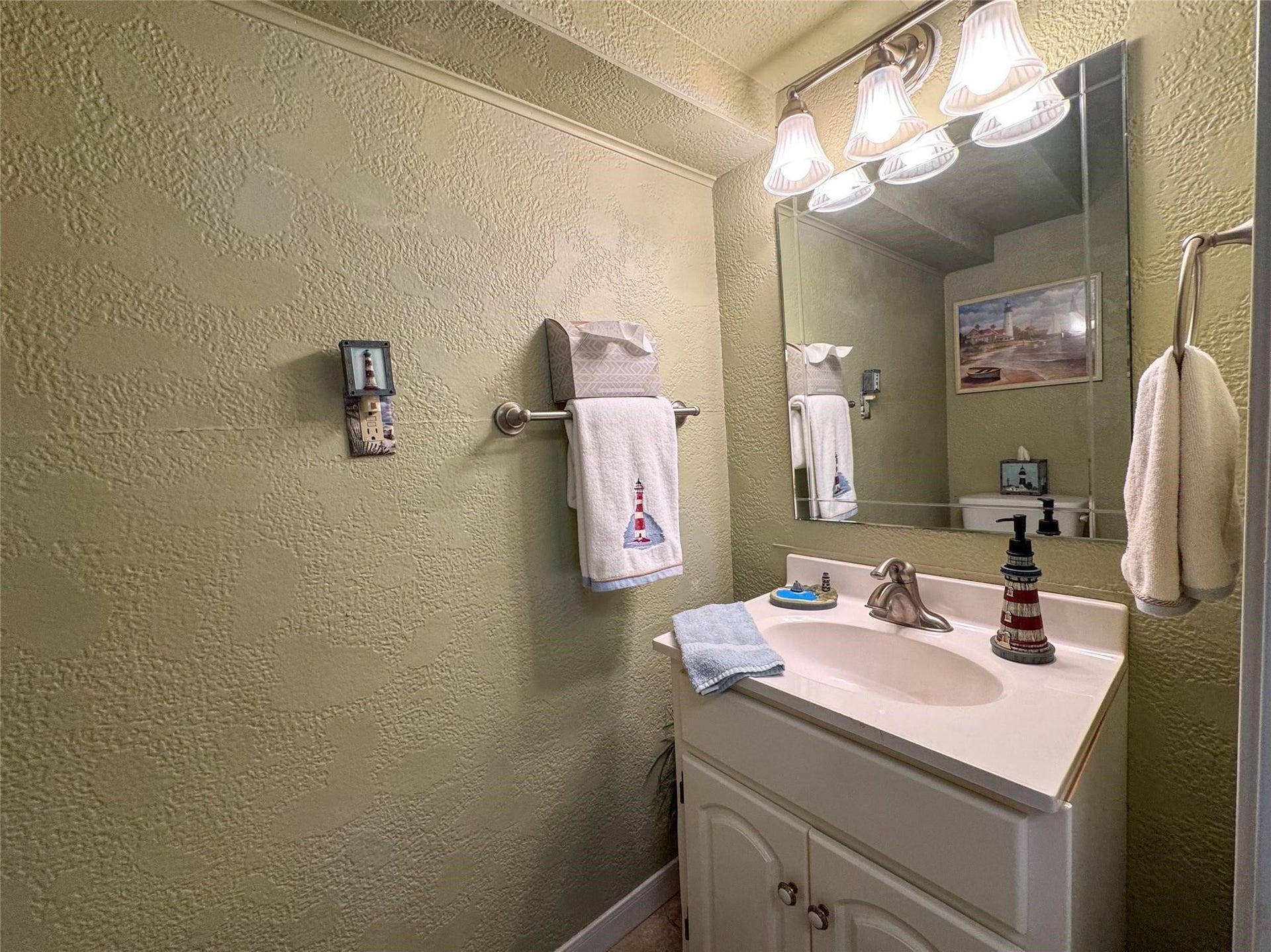 1031 Redfish Street, Bayou Vista, TX 77563 This bathroom features textured green walls, a large mirror with overhead lighting, and a white vanity. Nautical-themed accents add charm, including lighthouse decor and towels.