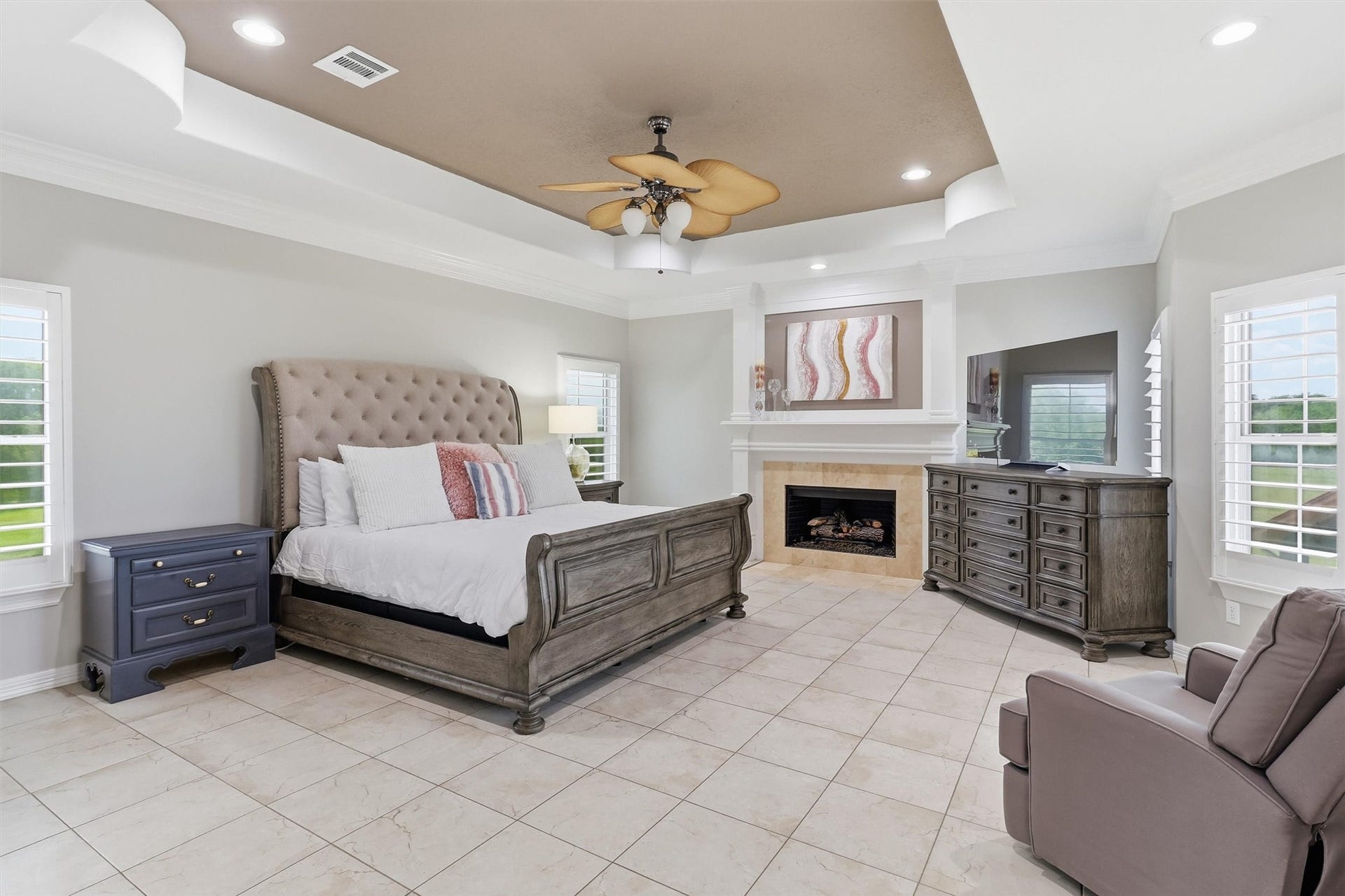 This luxurious primary bedroom offers a tray ceiling, a cozy fireplace, and abundant natural light from surrounding windows. Tile flooring and generous space for a sitting area make this a true private retreat.