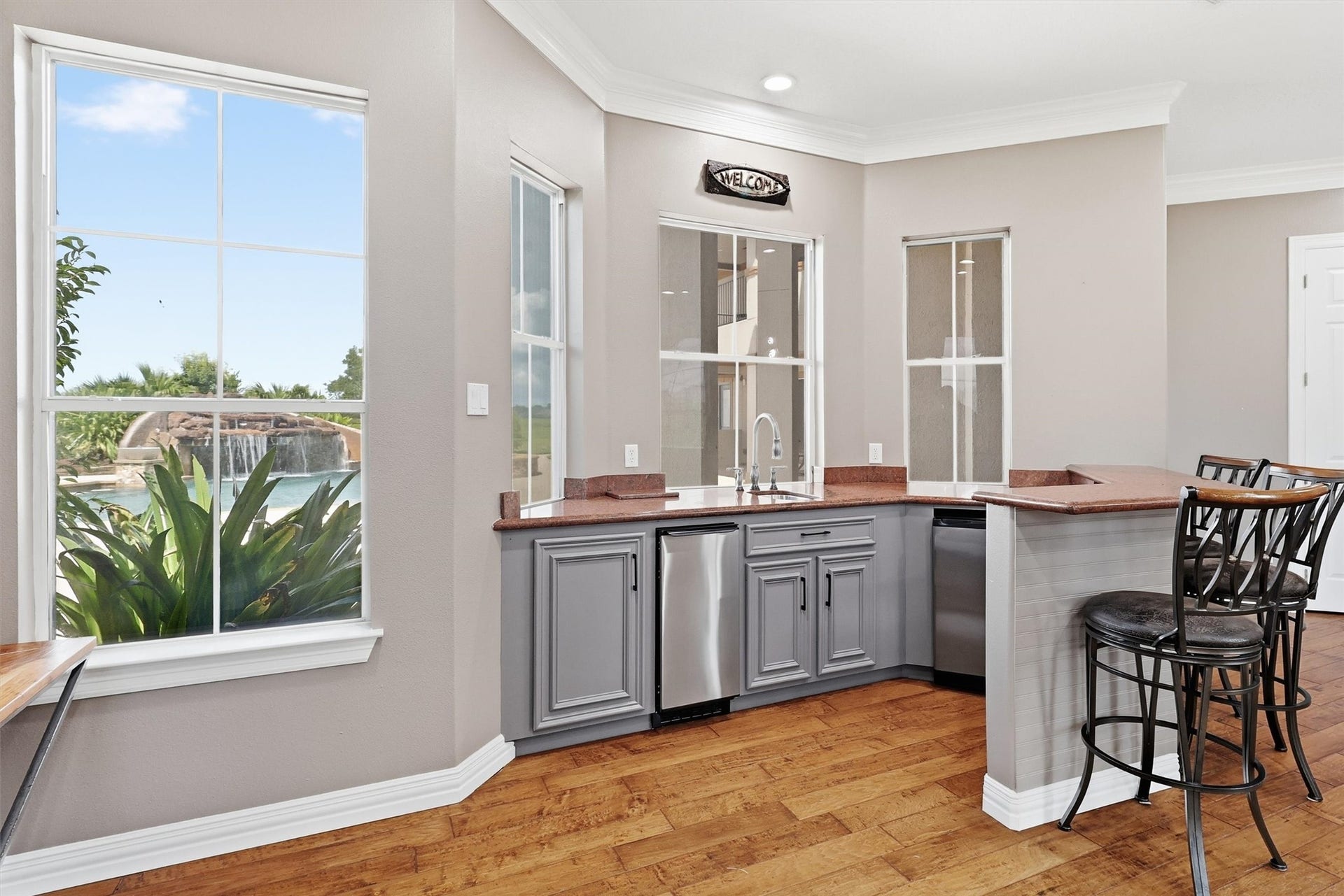 This handy wet bar area features stone countertops, a stainless beverage fridge, and a built-in sink, all framed by charming windows overlooking the sparkling pool and waterfall feature just outside.