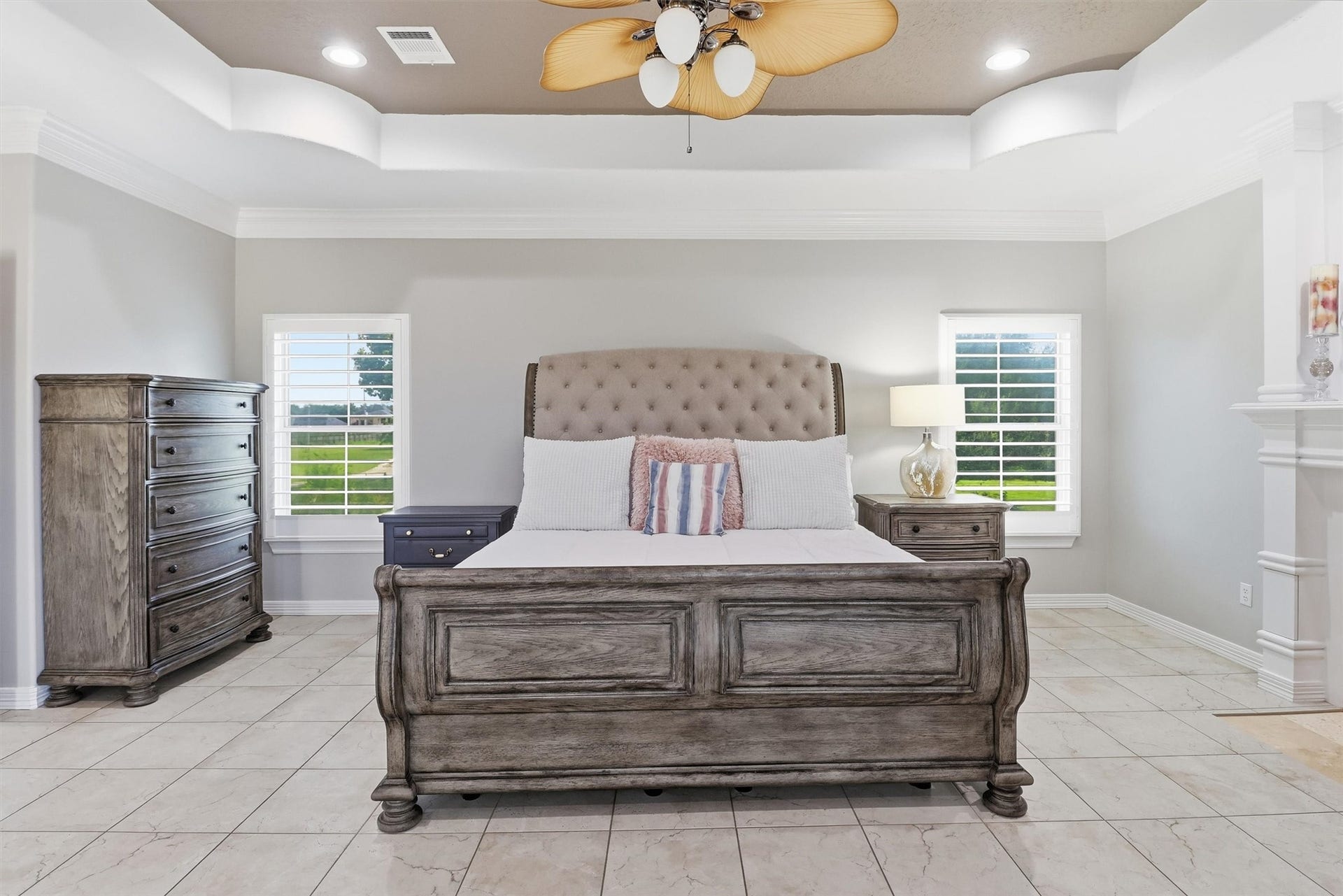 This luxurious primary bedroom offers a tray ceiling, a cozy fireplace, and abundant natural light from surrounding windows. Tile flooring and generous space for a sitting area make this a true private retreat.