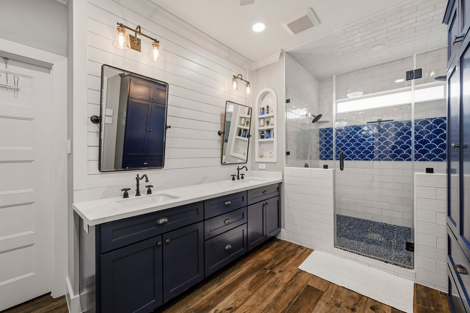 5702 Misty Island Court, Katy, TX 77494 Elegant bathroom featuring a double vanity with navy cabinets, shiplap walls, and a spacious glass-enclosed shower adorned with striking blue tile accents.