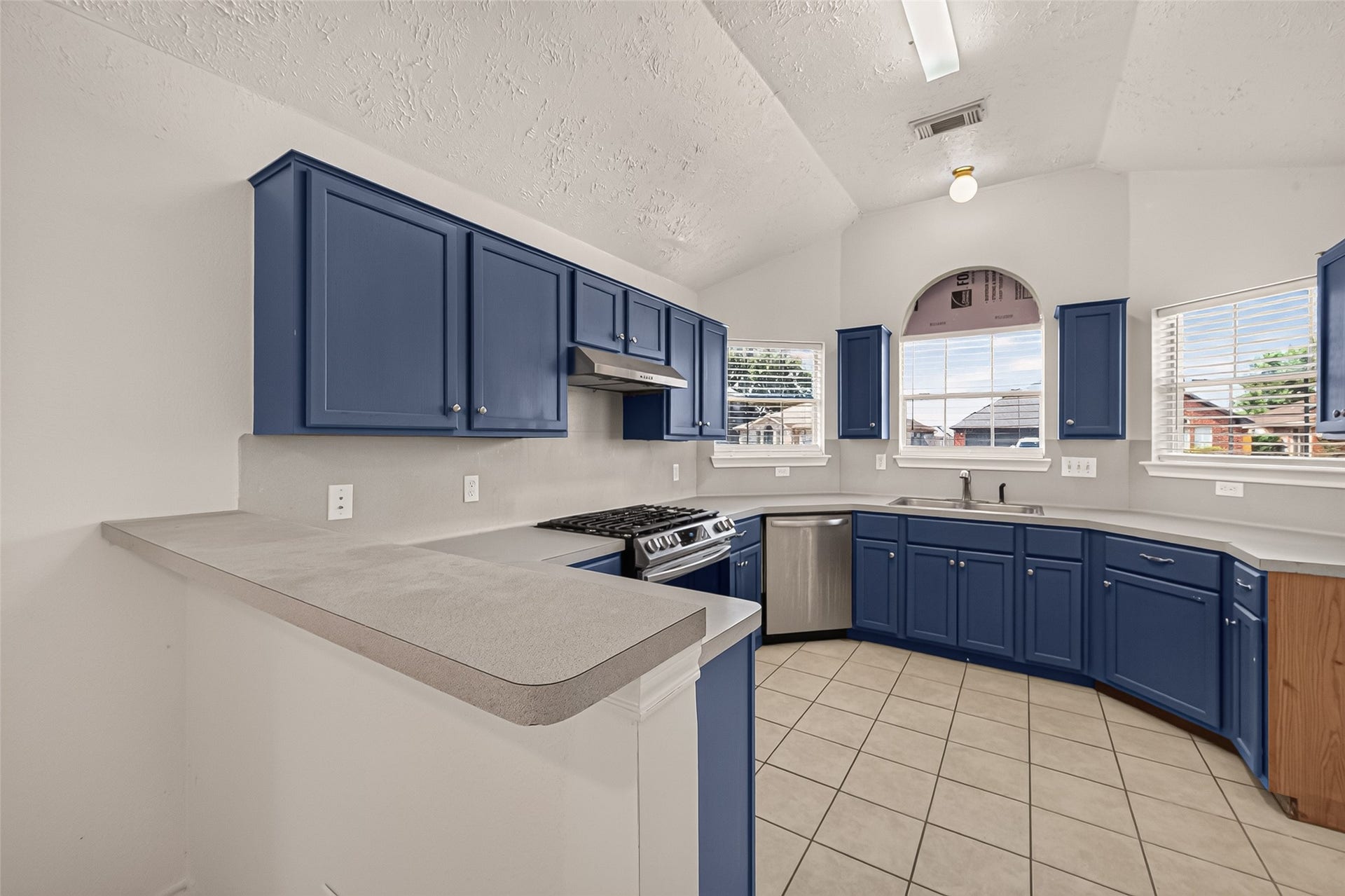 3356 Rachel Lane, Katy, TX 77493 This U-shaped kitchen has navy blue shaker cabinets, gray laminate countertops with a breakfast bar, stainless steel appliances, beige tile floors, and a vaulted textured ceiling with plenty of natural light.
