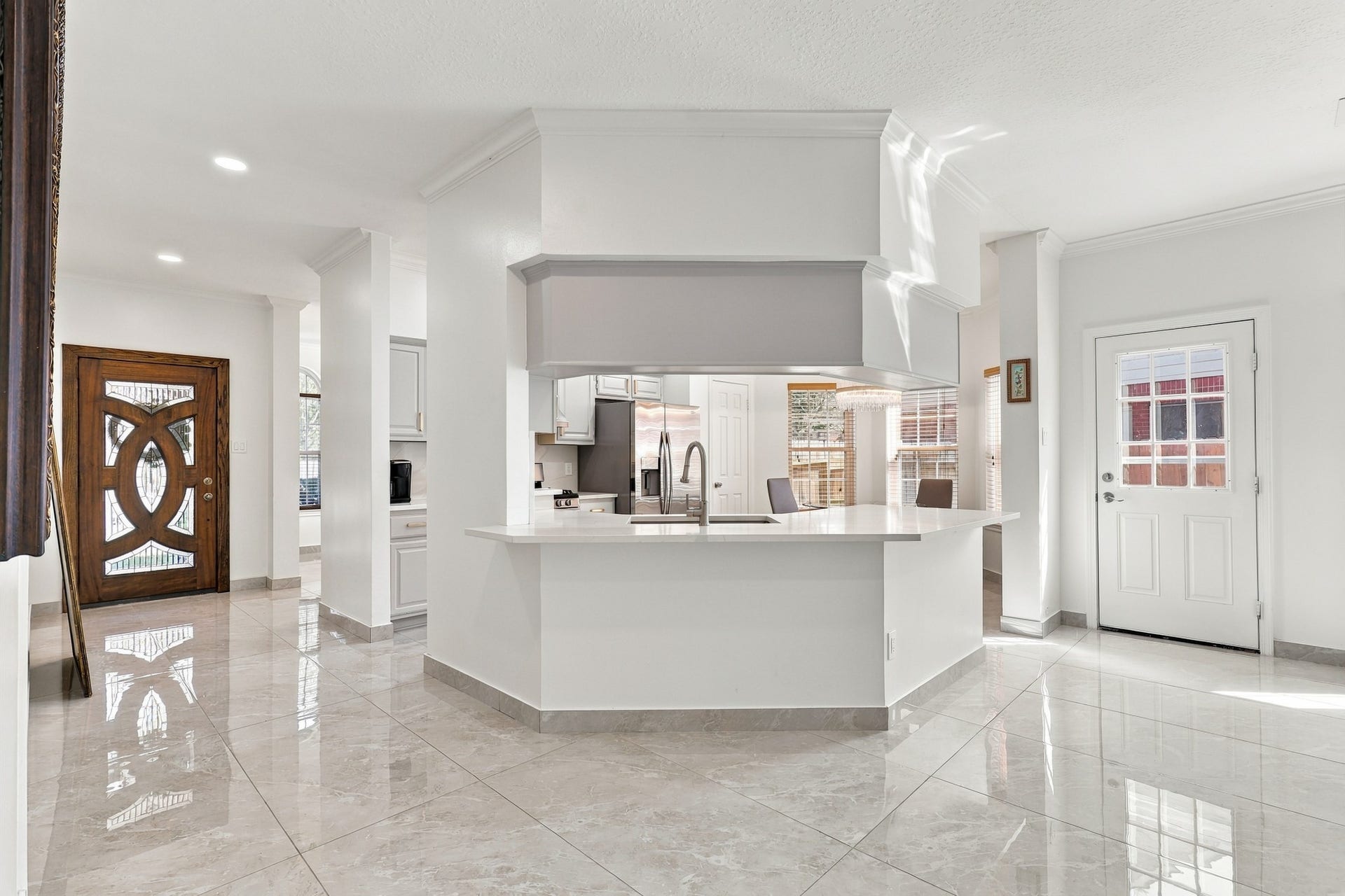 14538 Circlewood Way, Houston, TX 77062 Bright, open-concept layout featuring a spacious kitchen with wraparound breakfast bar, crisp white countertops, and views into the adjoining living and dining areas. Soaring ceilings, decorative columns, crown molding, recessed lighting, and polished large-format tile flooring enhance the home's airy feel, while the distinctive wood entry door adds architectural character.