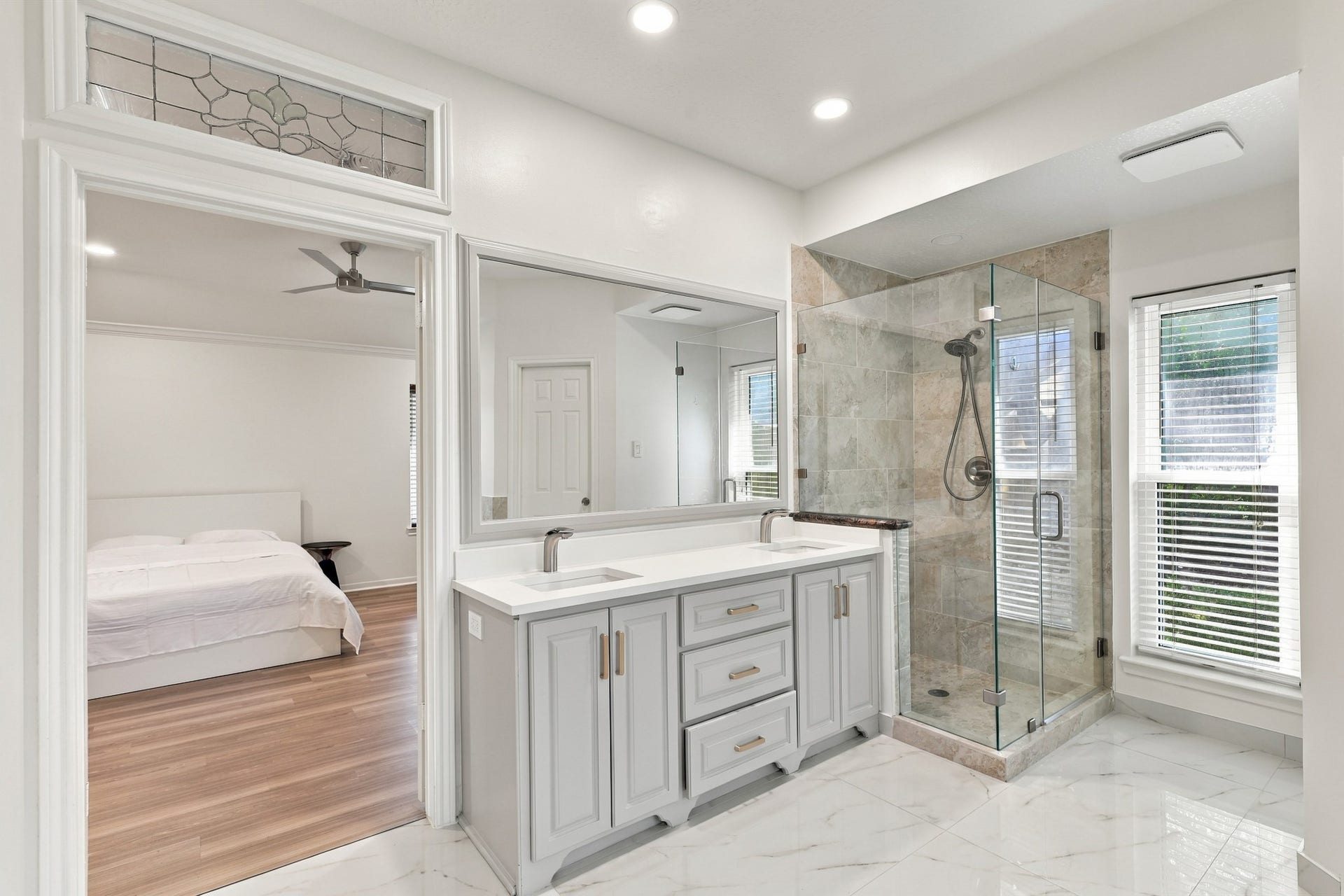14538 Circlewood Way, Houston, TX 77062 Beautifully appointed primary bathroom featuring a double-sink vanity with generous cabinetry, sleek countertops, and brushed gold hardware. A spacious glass-enclosed shower with floor-to-ceiling tile, elegant marble flooring, and natural light from the large window complete the space.