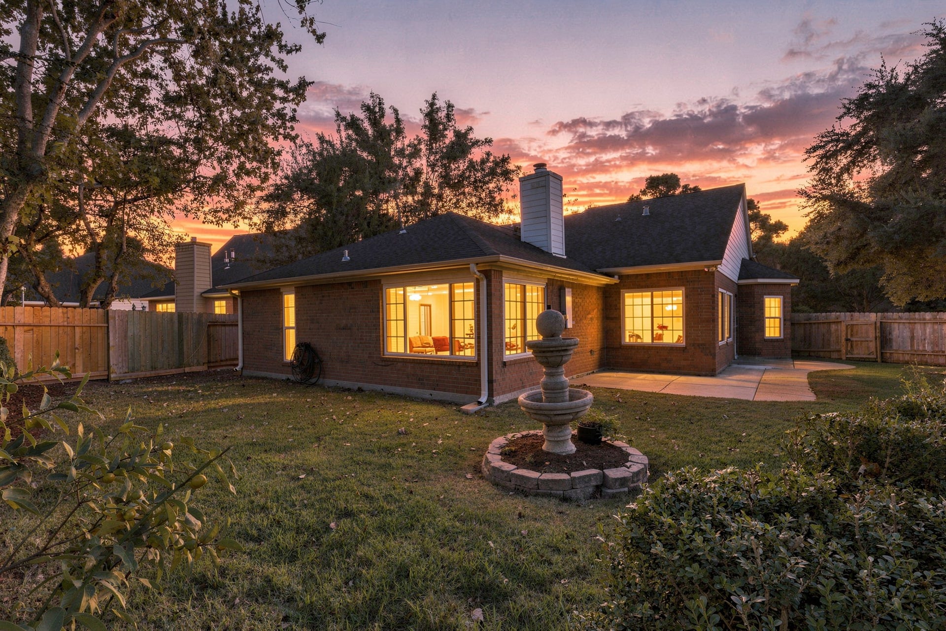 14538 Circlewood Way, Houston, TX 77062 As the sun sets, this beautiful brick home takes on an inviting glow, with warm interior lighting shining through its expansive windows. The backyard features a spacious patio, mature trees, a decorative tiered fountain, and a fully fenced lawn. A charming setting for relaxing evenings, outdoor gatherings, and enjoying the comforts of home.