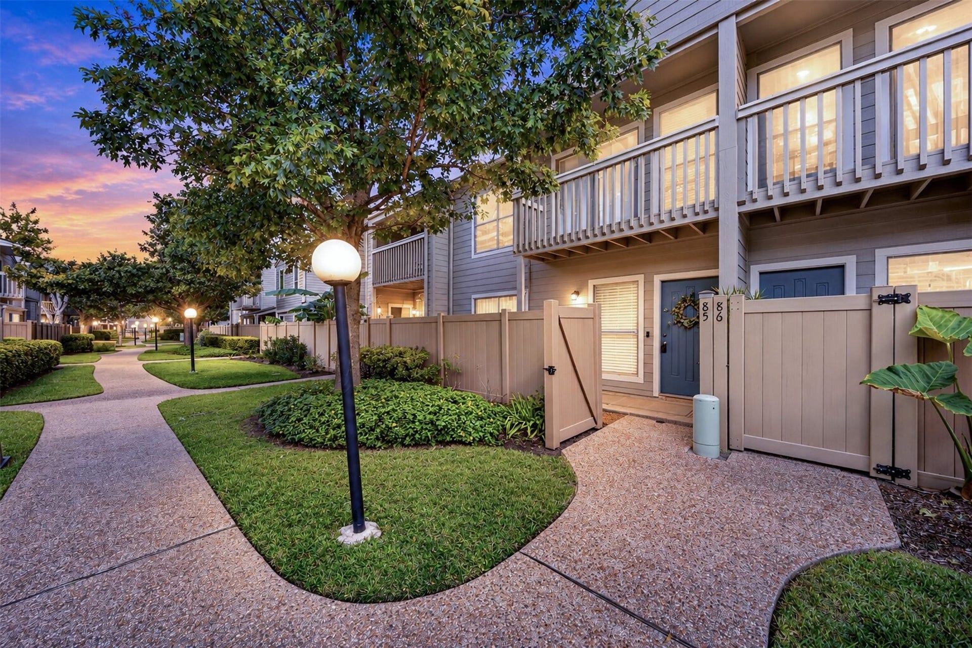 2379 Briarwest Boulevard, #85, Houston, TX 77077 Twilight settles over Unit 85 as warm light glows from the windows and balcony. The gated entry, tree-lined walkways, and lampposts give the community an evening glow.