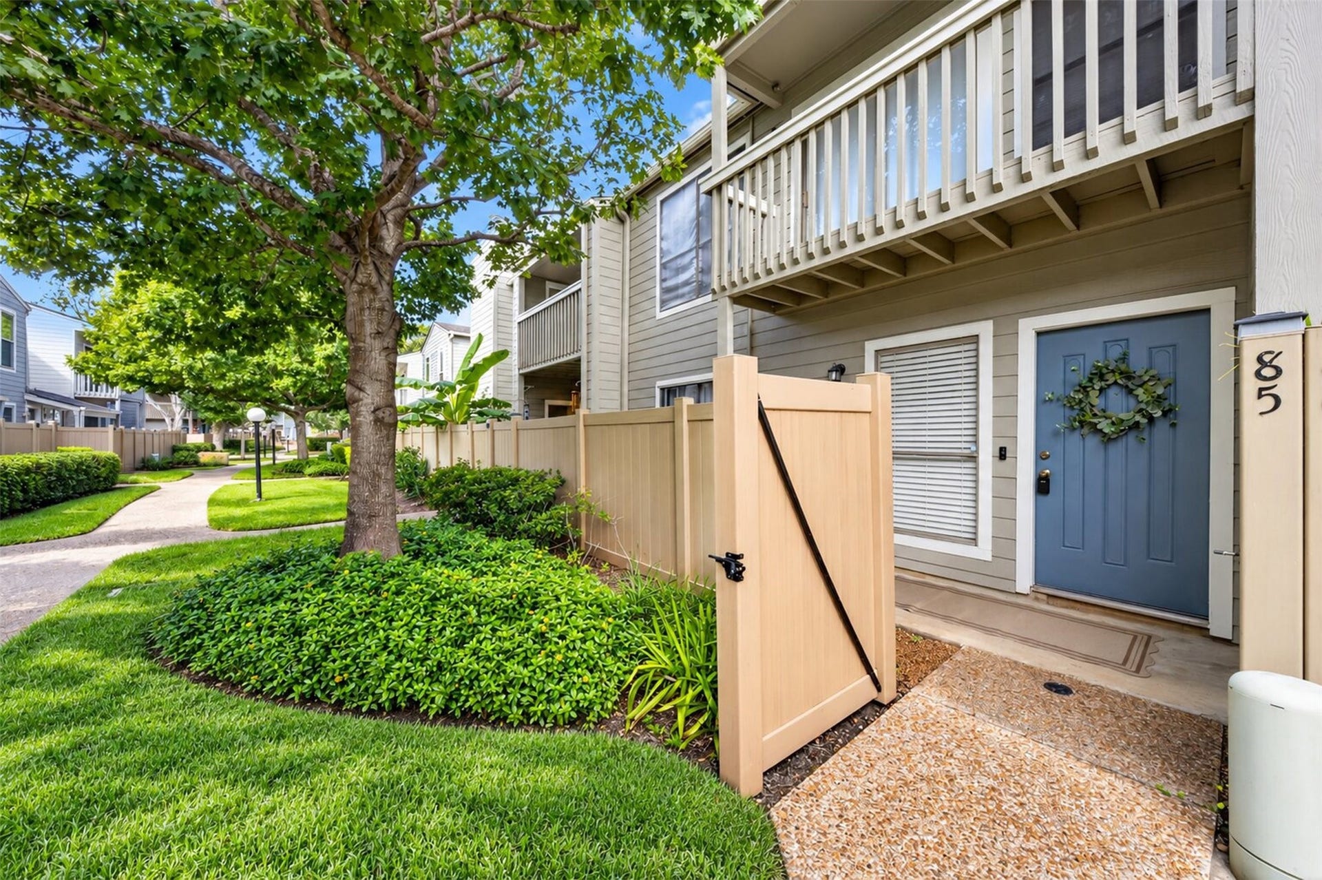 2379 Briarwest Boulevard, #85, Houston, TX 77077 Welcome home to Unit 85: a slate-blue front door with a greenery wreath, a gated front patio, and a second-story balcony, all shaded by mature trees in a manicured, walkable community.