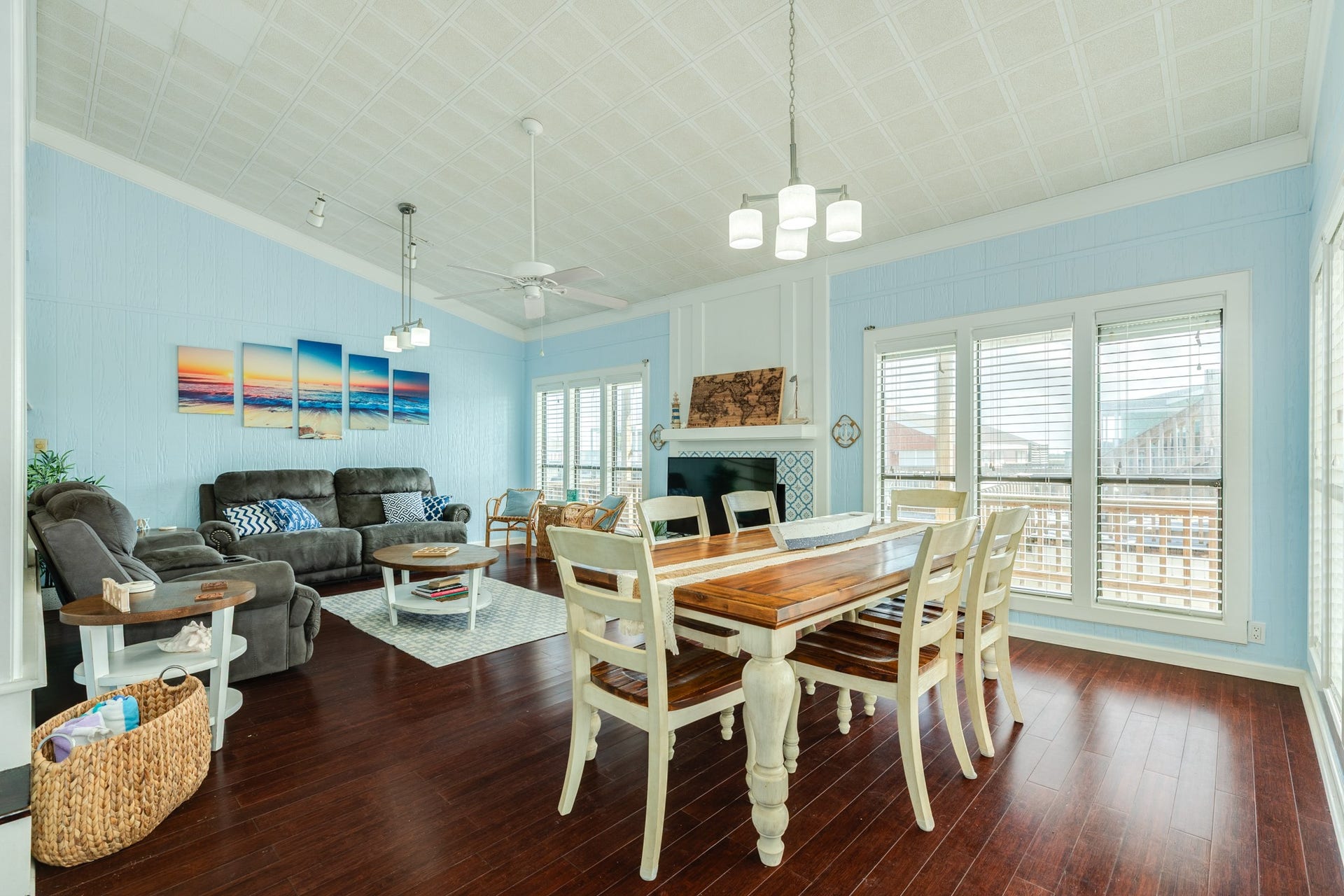 Bright, open-concept living and dining space features vaulted ceilings, rich wood-look flooring, coastal blue walls, decorative lighting, and abundant natural light from multiple windows.