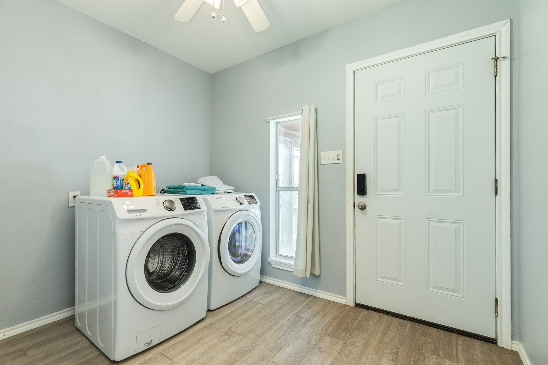 Dedicated laundry area features a side-by-side front-loading washer and dryer, light wood-look flooring, and a window for natural light. Convenient exterior door provides direct access to the outdoor area.