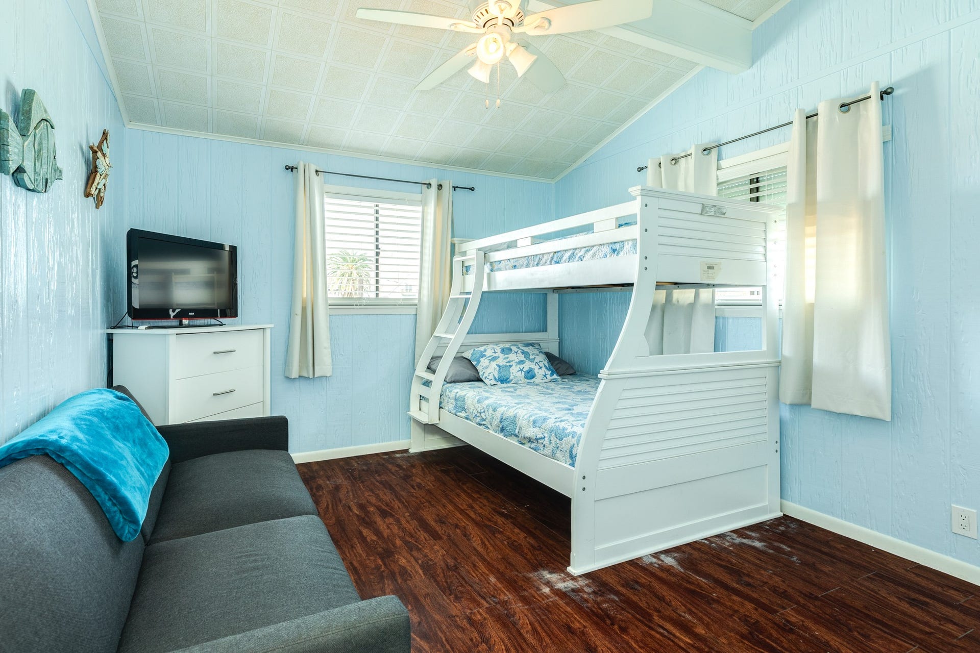 Coastal-inspired bedroom features a white bunk bed, additional sofa seating, vaulted ceilings, and dark wood-look flooring, offering flexible sleeping and lounging space.