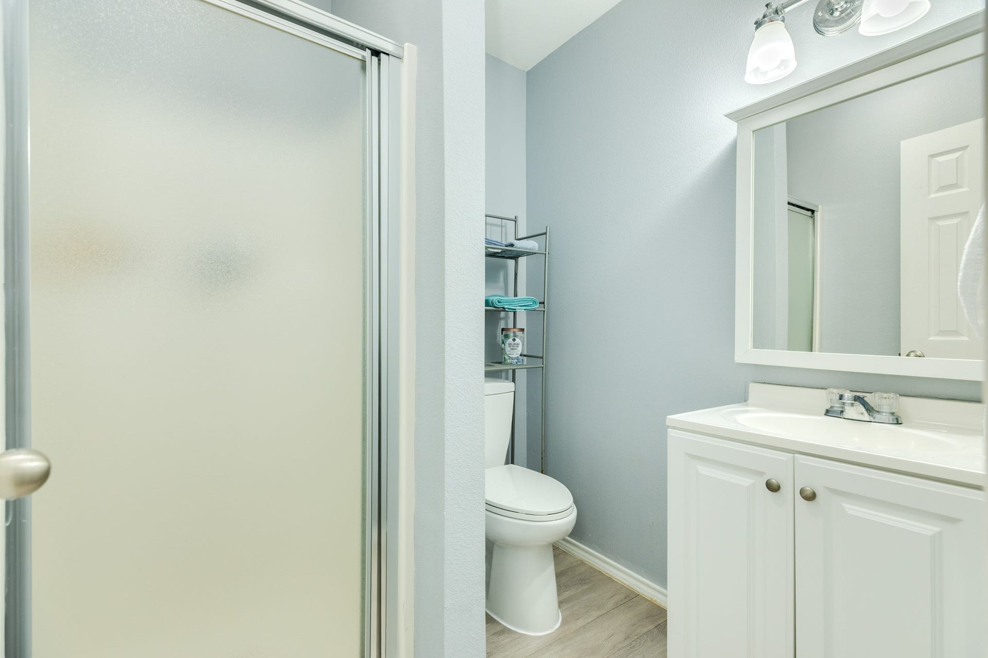 Bright bathroom features a white vanity with cabinet storage, framed mirror, contemporary lighting, and a glass-enclosed shower with frosted doors. Neutral finishes and wood-look flooring create a clean, cohesive design. Ground floor.
