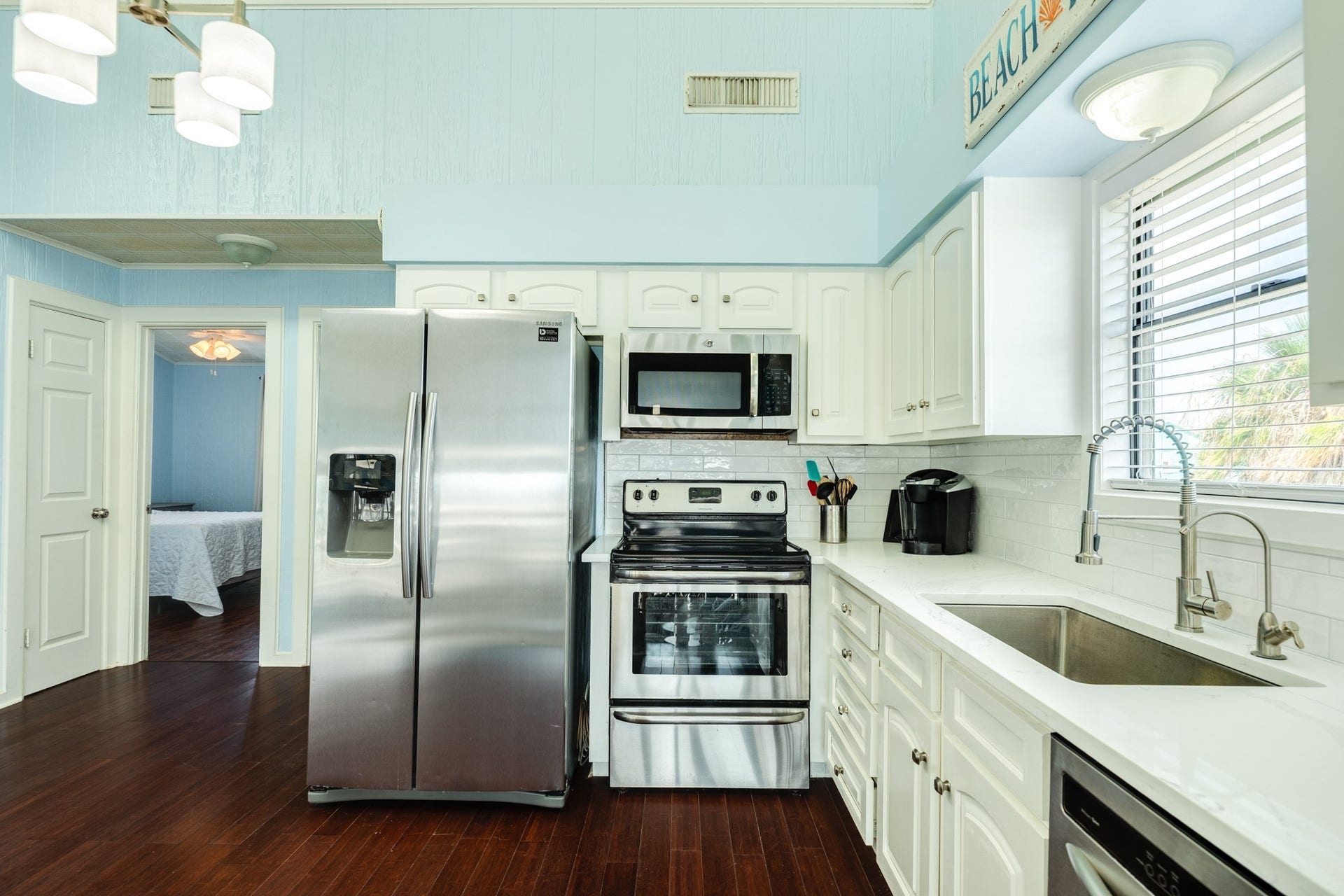Kitchen features stainless steel appliances, white cabinetry, light countertops, subway tile backsplash, and a deep single-basin sink with a spring-style faucet beneath a window.