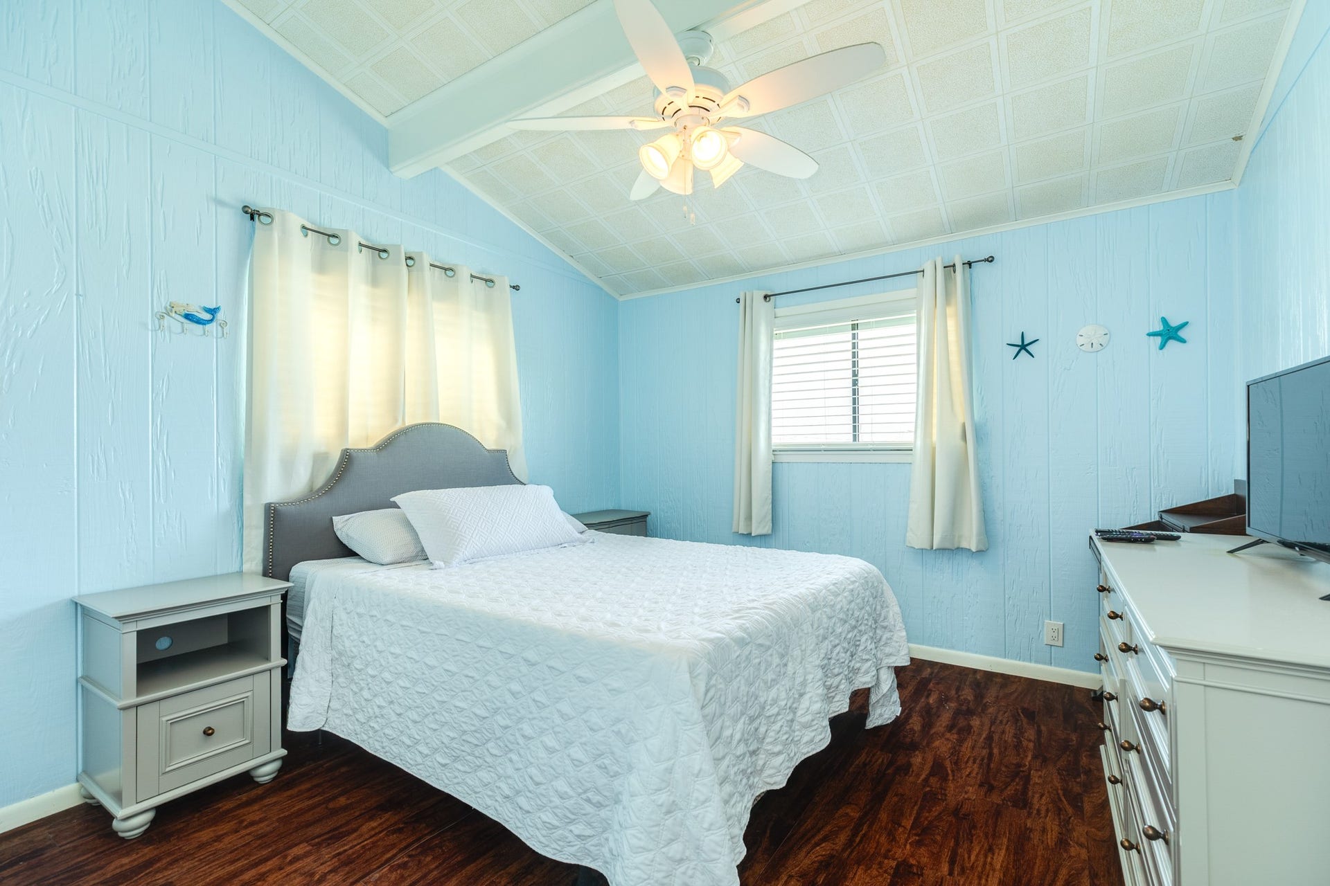 Comfortable bedroom features soft blue walls, vaulted ceilings, dark wood-look flooring, a ceiling fan, and windows with white curtains that welcome natural light.