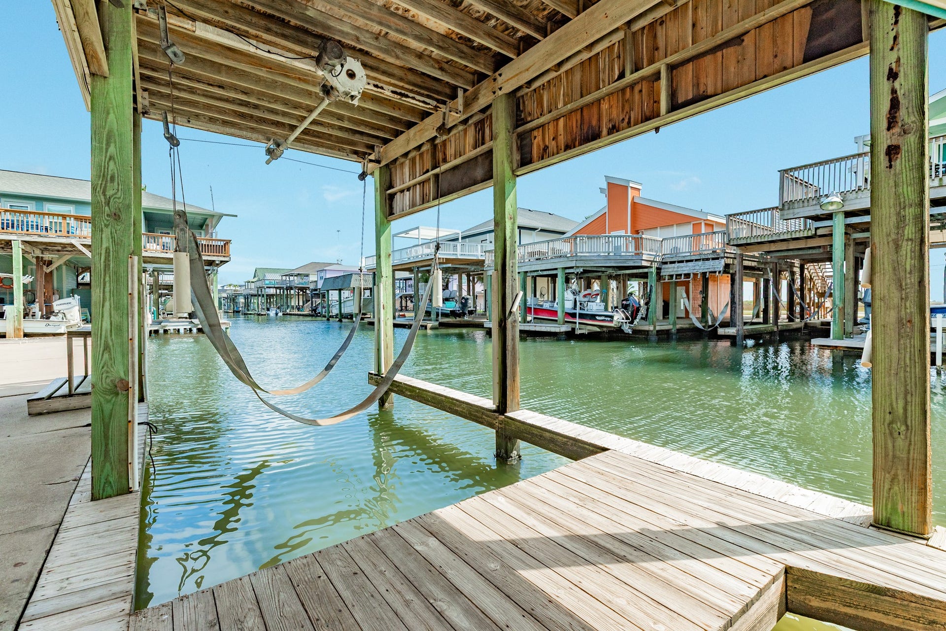 Another view of the covered boat lift showcases the overhead lifting equipment, wooden dock, and canal extending through the waterfront neighborhood.