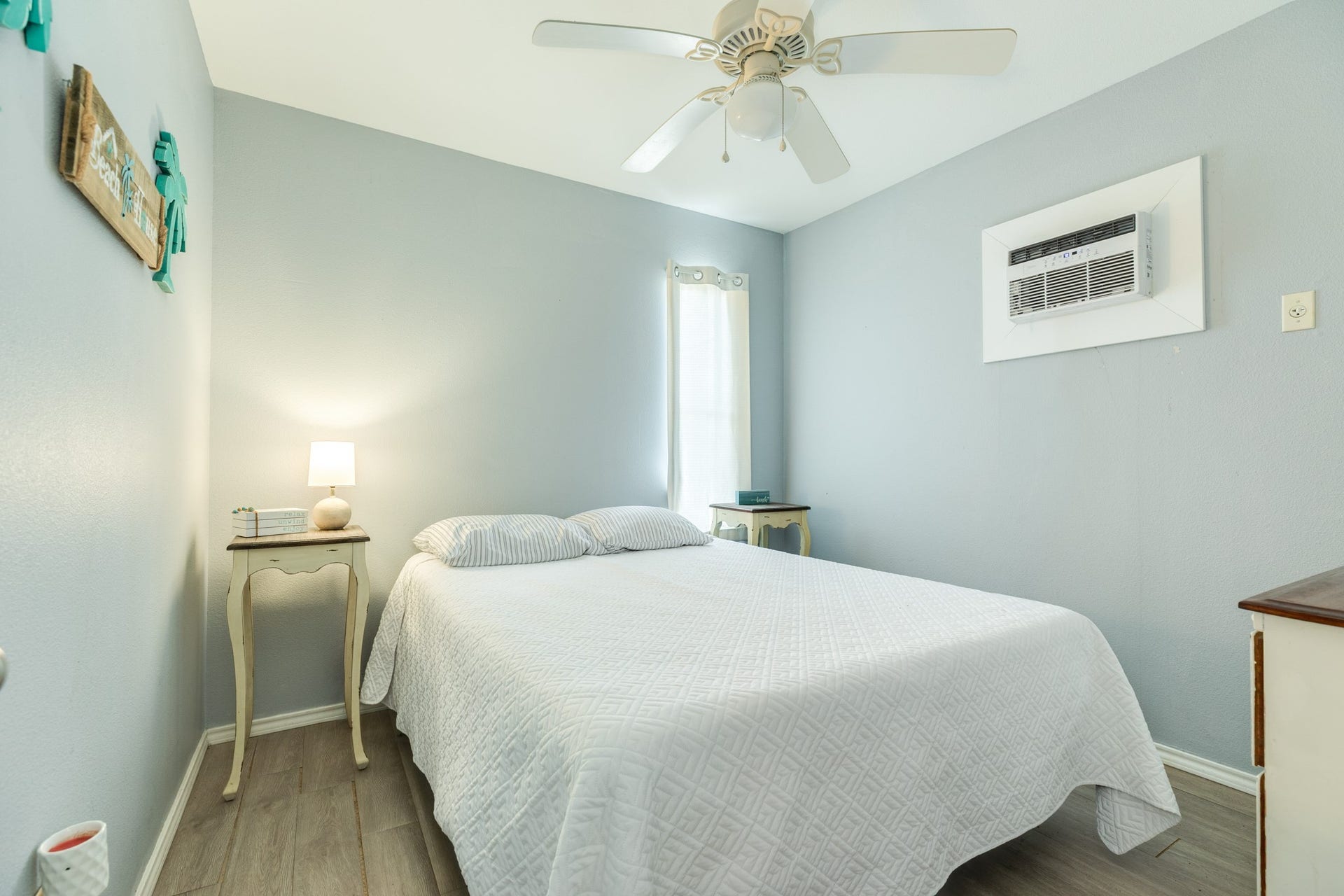 Inviting ground floor bedroom features soft gray-blue walls, light wood-look flooring, a ceiling fan, and a window providing natural light. A wall-mounted air conditioning unit offers additional comfort.