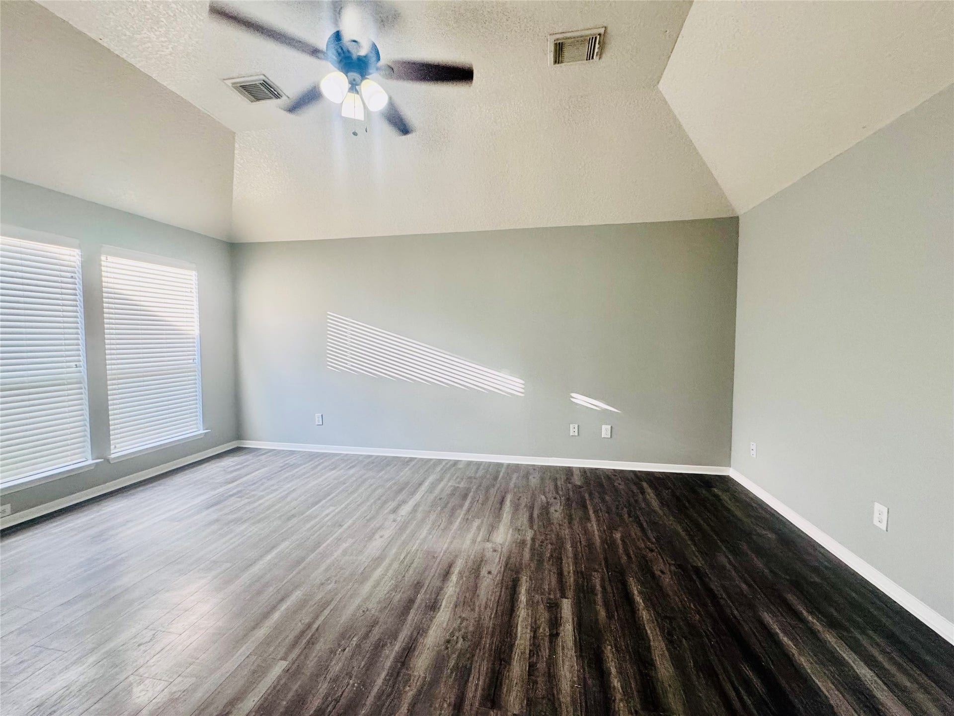 Spacious primary bedroom with vaulted ceilings, elegant wood flooring, and abundant natural light through large windows, featuring a modern ceiling fan for comfort.