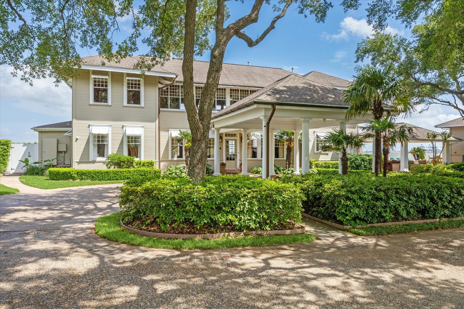 This historic two-story home features a classic design with large windows and an inviting front porch. It is surrounded by lush landscaping, including mature trees and well-maintained hedges, offering a serene and private setting. The driveway provides convenient access and adds to the overall curb appeal.