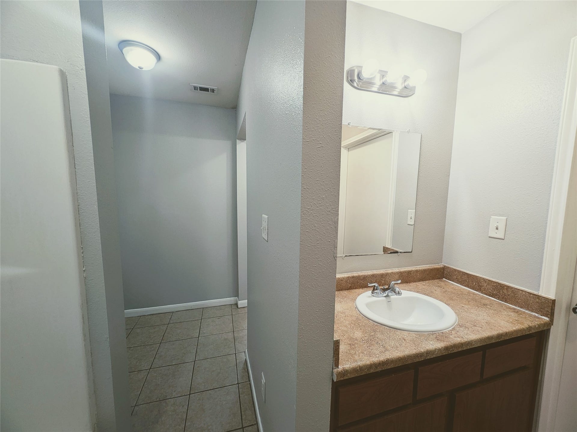 This photo showcases a simple bathroom with a single sink vanity featuring a brown countertop, wooden cabinetry, and a wall-mounted mirror. The space has tiled flooring and neutral-colored walls, creating a clean and modern look.