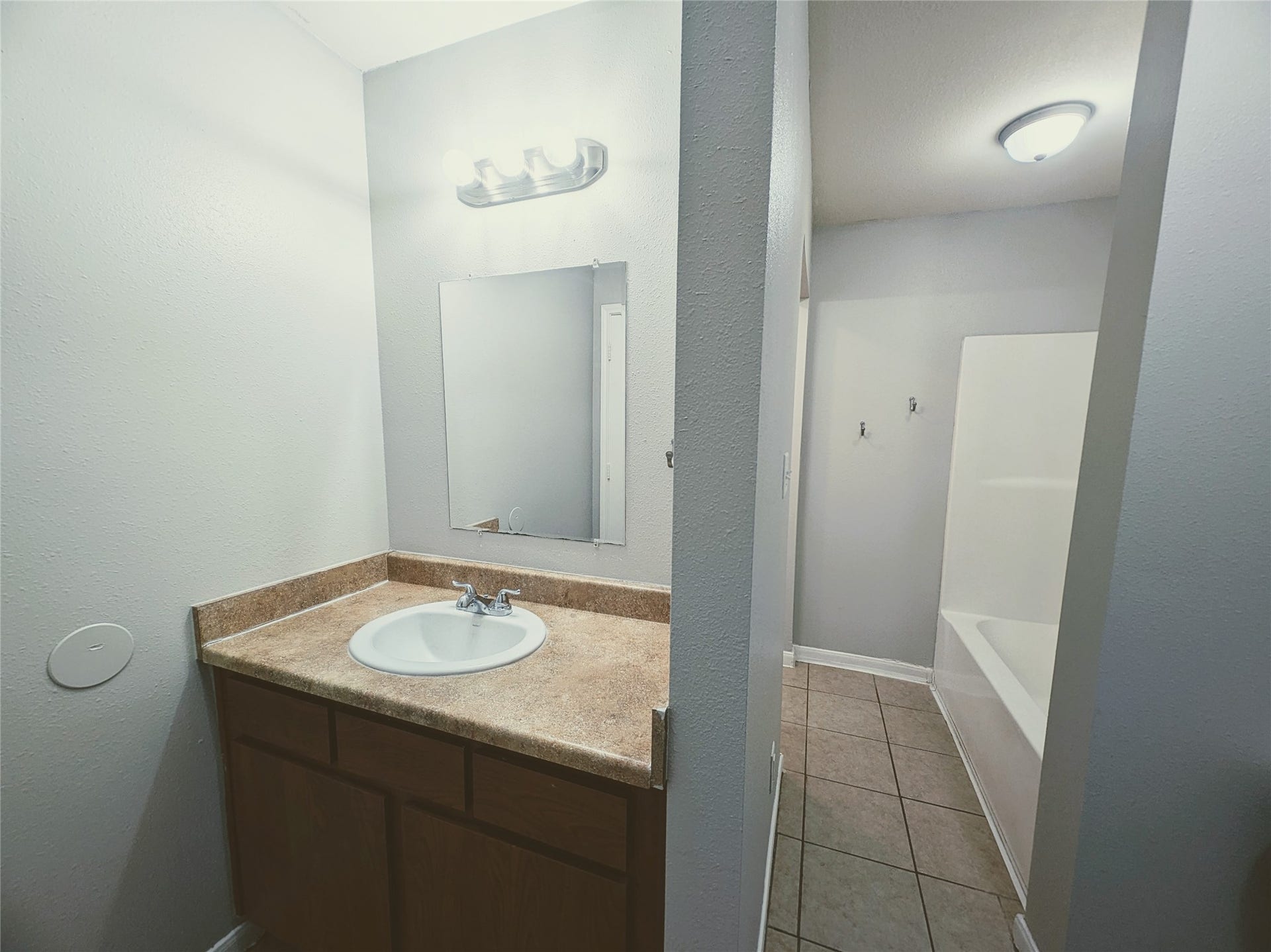 This photo shows a simple bathroom with a single sink vanity and a mirror. The countertop is a light brown, and the walls are painted a neutral color. The bathroom includes a bathtub and is well-lit with overhead lighting. The floor is tiled.