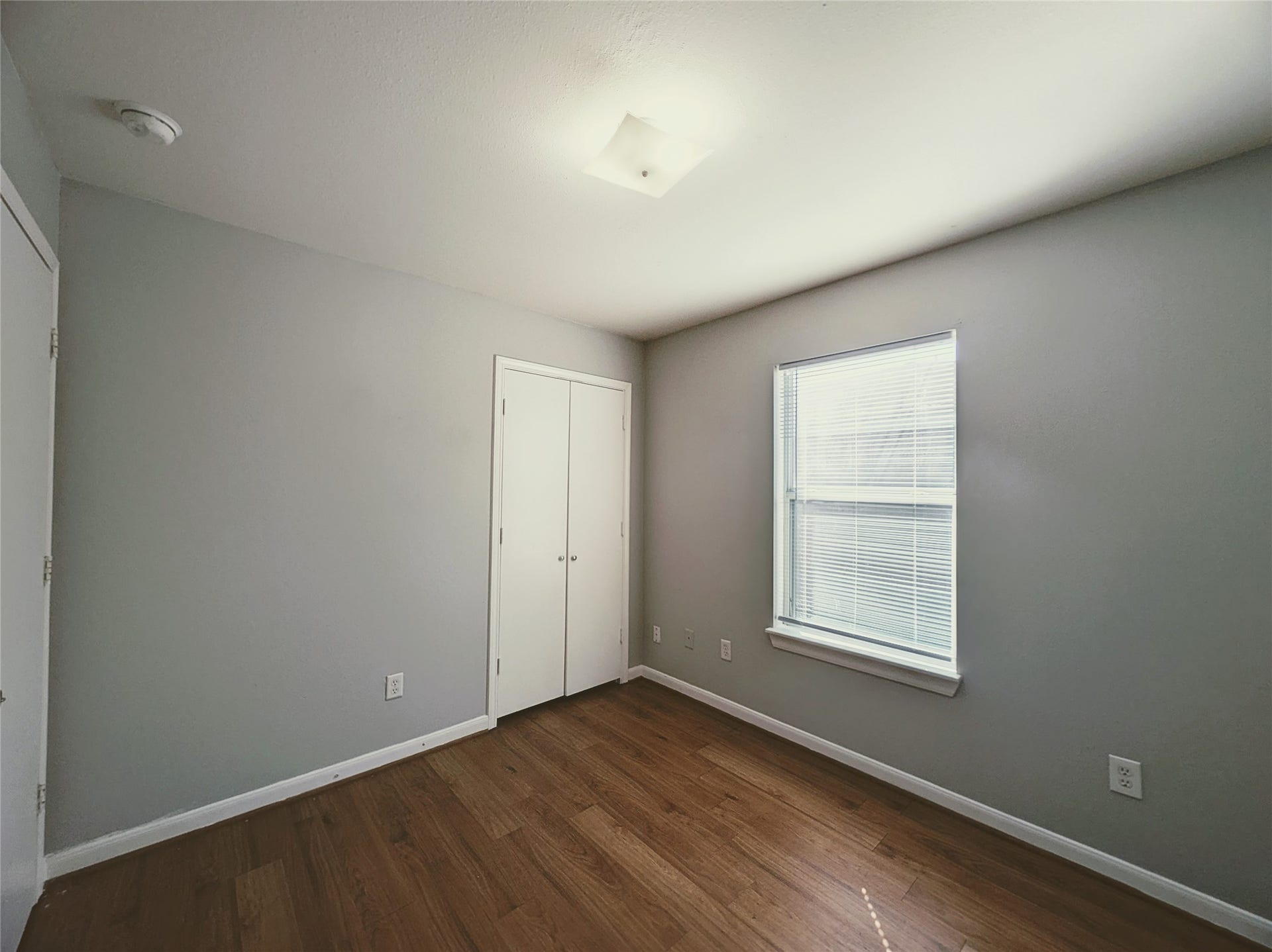 Bright room with hardwood floors, light gray walls, and a window with blinds. There's a small closet with double doors, and the ceiling features a modern light fixture.