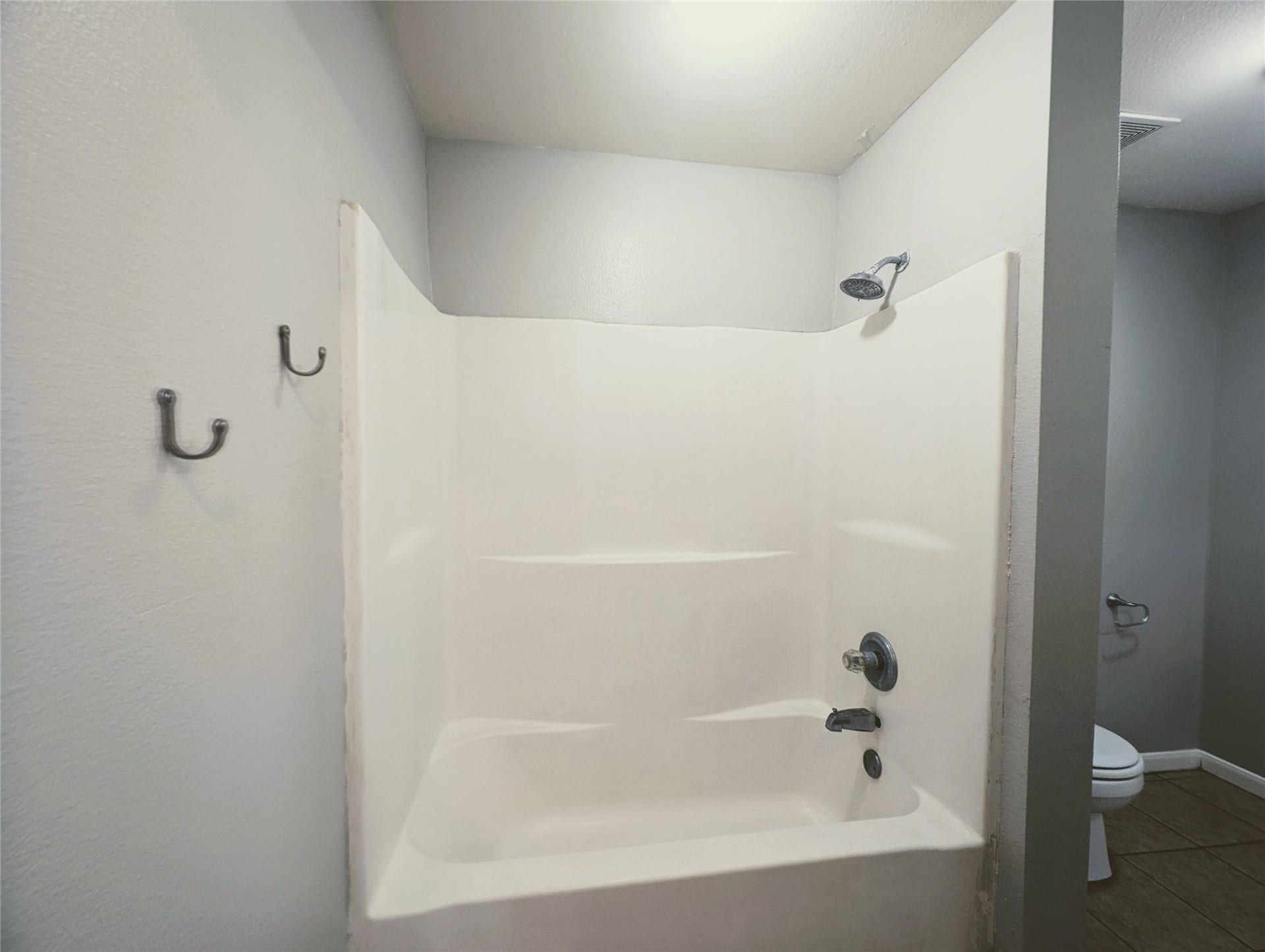 This photo shows a bathroom with a clean, white shower-tub combo, gray walls, and tiled flooring. There are two wall hooks and a visible toilet area, providing a functional and simple space.