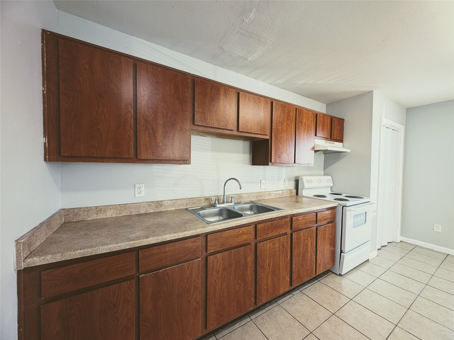 This kitchen features ample wooden cabinetry, a double sink, and an electric stove. The countertops are spacious, and the room is finished with tiled flooring, offering a clean and functional space.