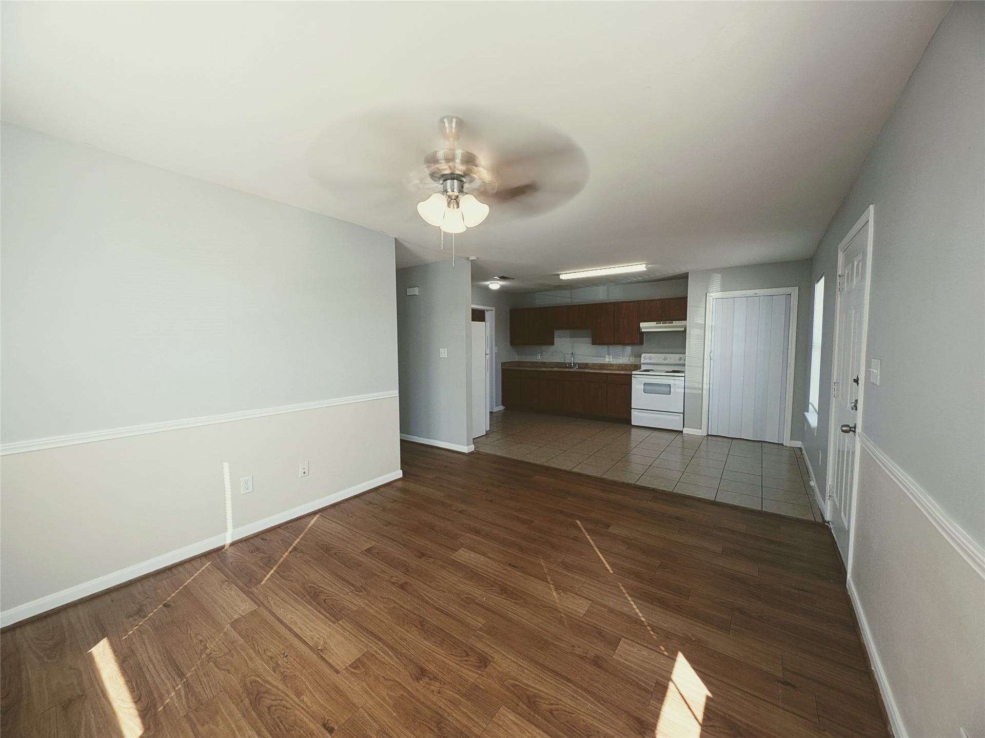 This photo shows a bright, open-concept living area with a ceiling fan and wood-like flooring. It seamlessly connects to a kitchen featuring tile flooring, wooden cabinets, and white appliances. The space is well-lit and has a clean, modern feel.