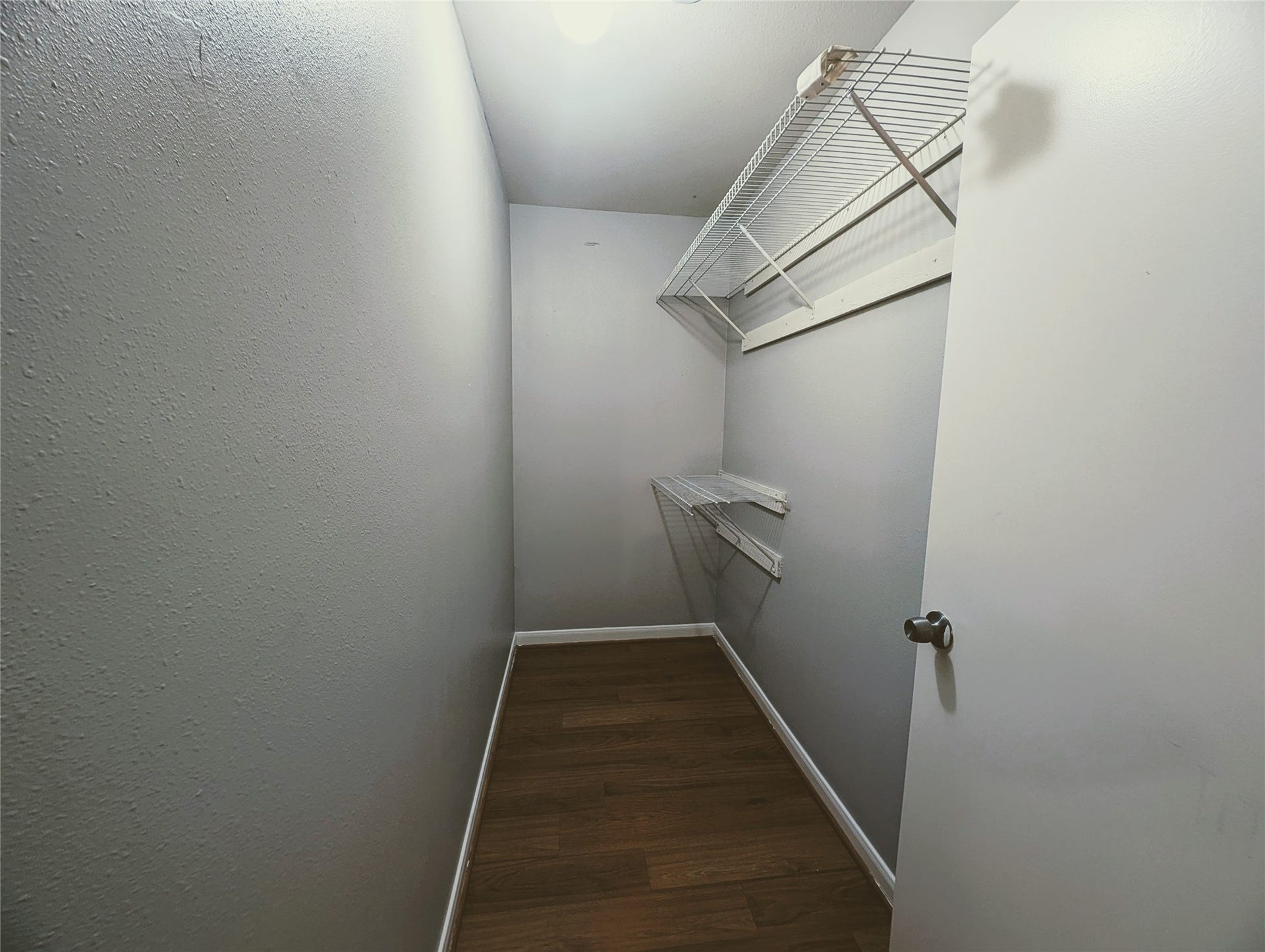 This photo shows a narrow walk-in closet with light gray walls, wooden flooring, and wire shelving units on one side for storage.