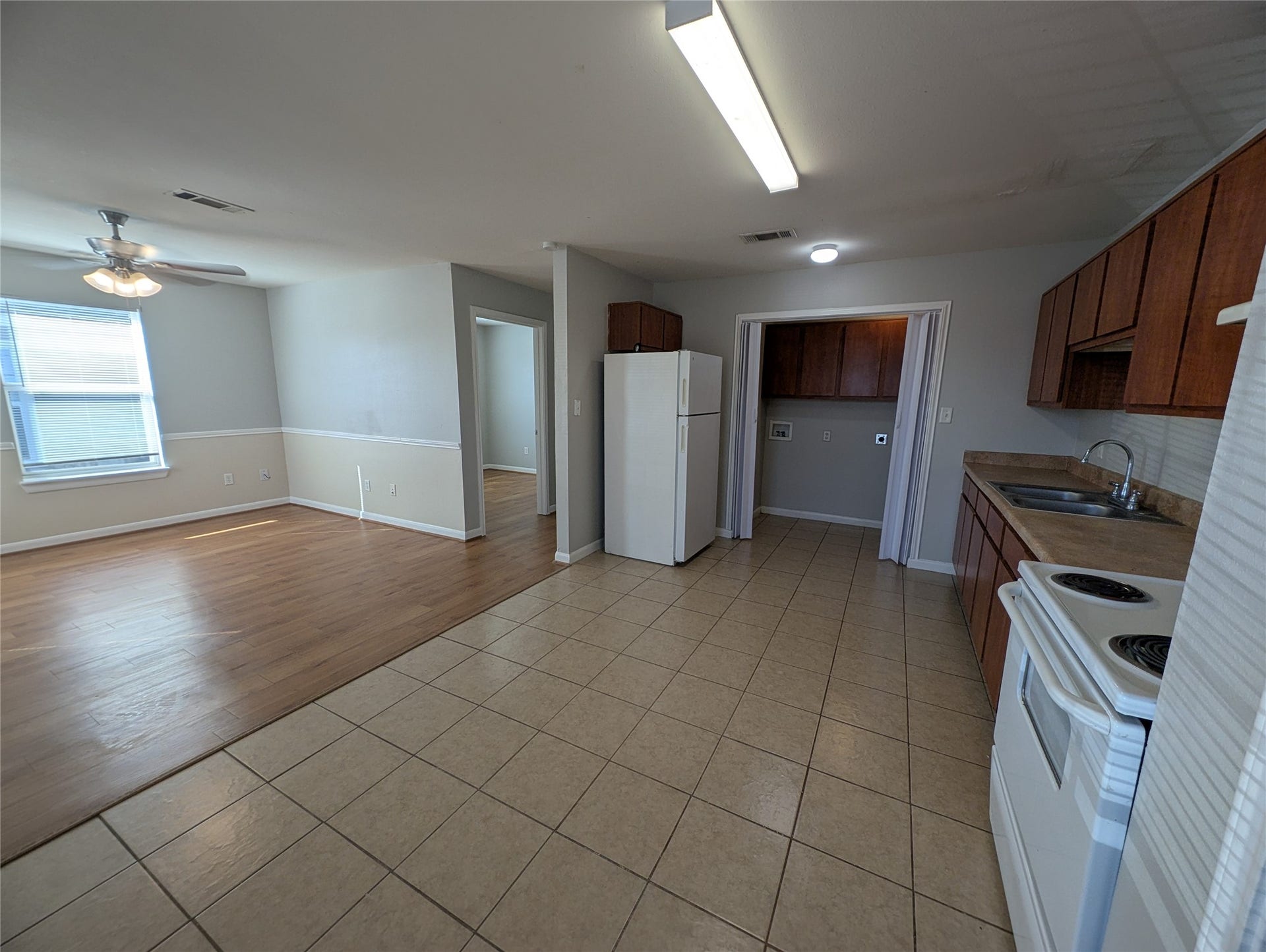 This photo shows an open-concept kitchen and living area. The kitchen has tiled floors, wooden cabinets, a white refrigerator, and a stove. Adjacent is a living space with wood flooring, a ceiling fan, and natural light from a window. There's a doorway leading to another room.