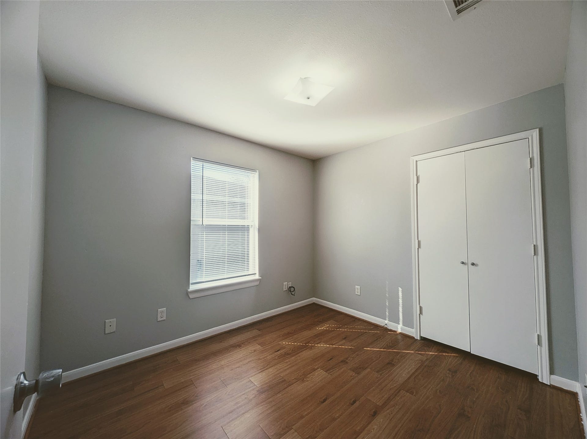 This photo shows a bright, compact bedroom with light gray walls, a single window, and a double-door closet. The room features wood-look flooring and a modern ceiling light, providing a clean, versatile space.