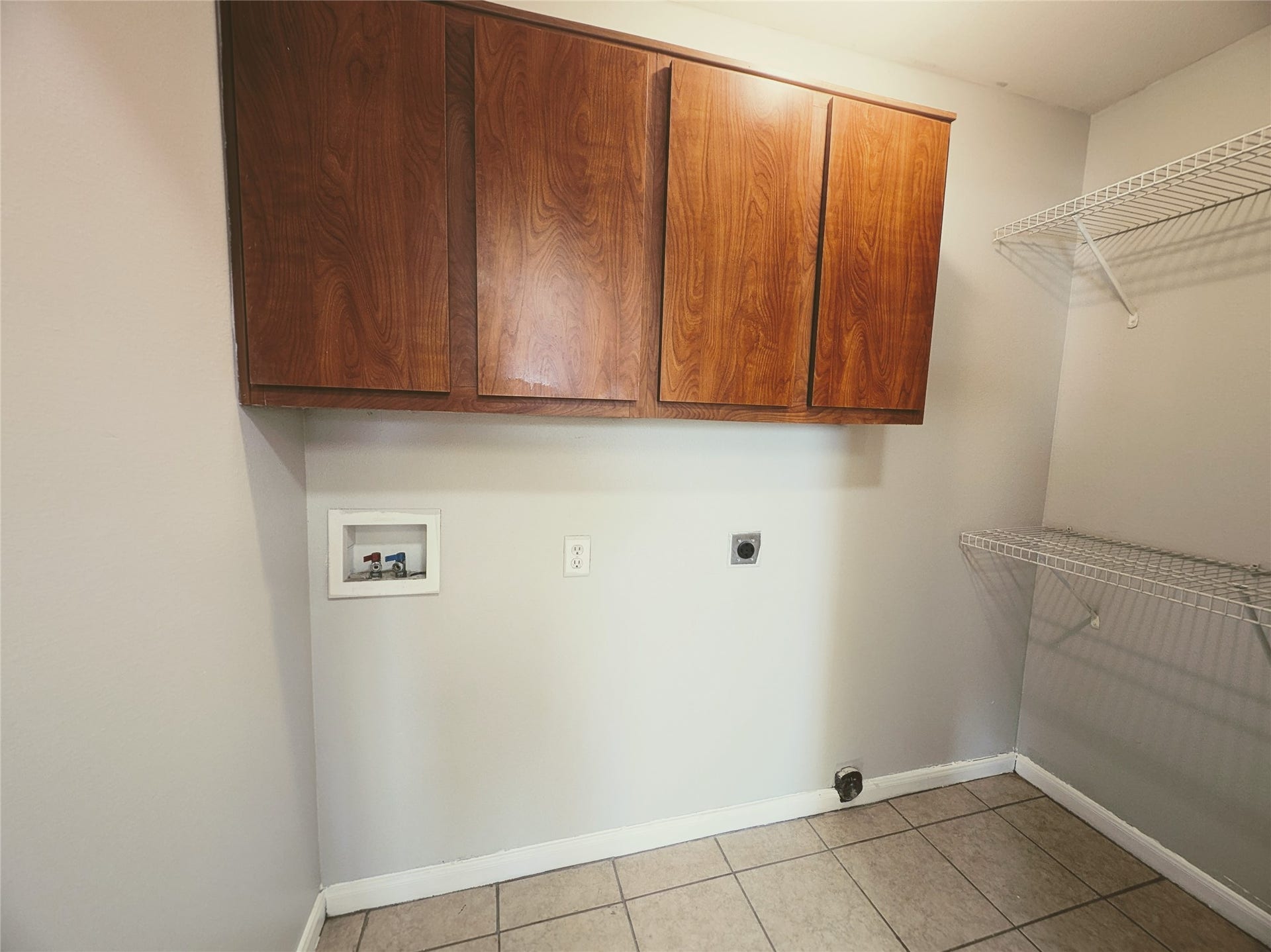 This photo shows a utility or laundry room with tiled flooring, wooden cabinets for storage, and wire shelving. The space is ready for appliance installation with necessary connections visible.