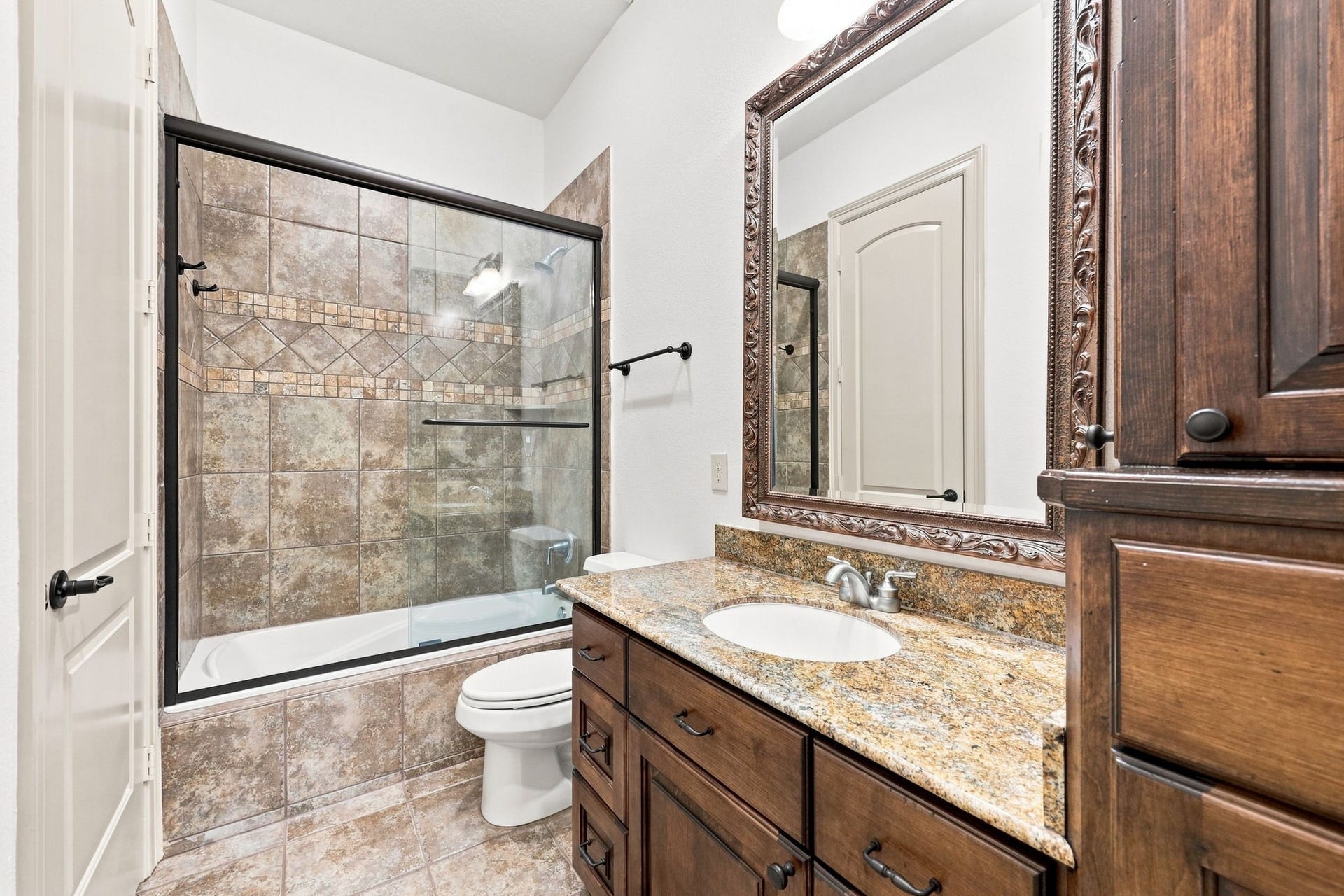 5411 Woodbury Street, Fulshear, TX 77441 This Full Bath serves the two Secondary Bedrooms featured in the previous 2 photos. The bath offers a tub and shower combination, custom cabinetry, a gorgeous granite countertop, and beautiful tilework, creating a stylish and functional space for family and guests.
