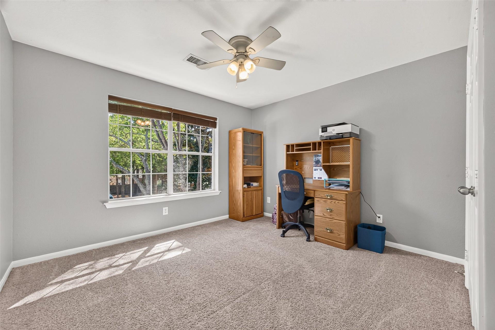 Spacious upstairs secondary bedroom featuring a large window overlooking mature trees, ceiling fan, neutral carpeting, and ample space for a bedroom or home office.