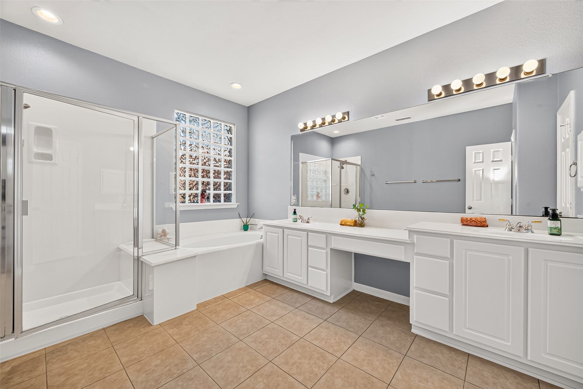 Spacious primary bathroom featuring dual vanities, a relaxing soaking tub, separate glass-enclosed shower, and glass-block window providing natural light.