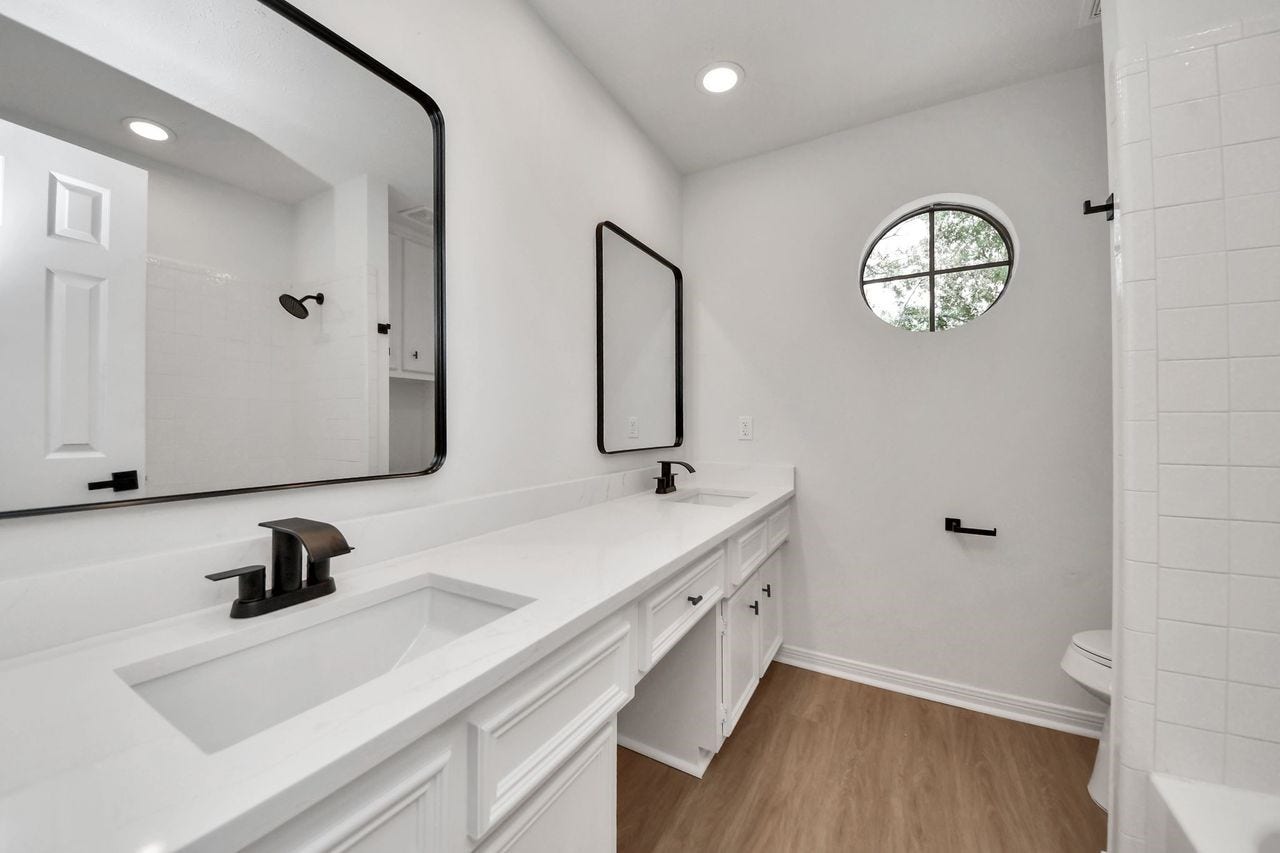 15047 Tramore Drive, Houston, TX 77083 This beautifully refreshed full bathroom features an extended dual-sink vanity with quartz countertops, crisp white cabinetry, and contemporary black fixtures. A charming round window adds natural light and architectural character, while the tub and shower combination is finished with clean white tile.
