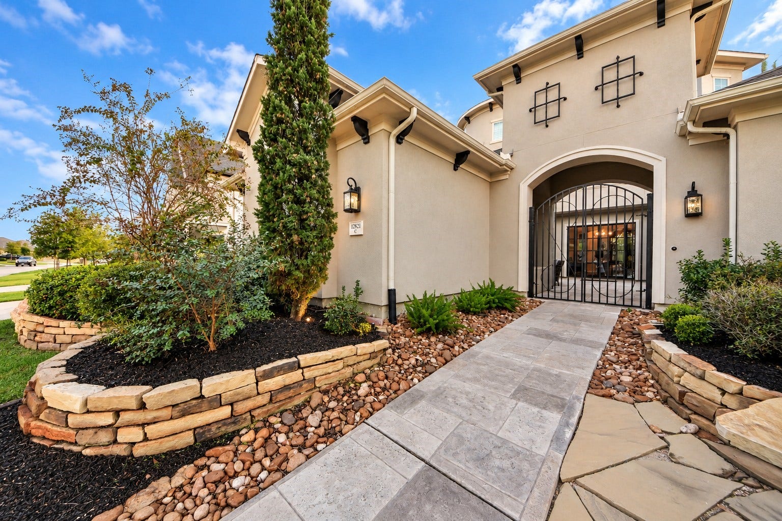 12821 N Palomino Lake Circle, Cypress, TX 77429 Inviting courtyard entrance featuring elegant double wrought-iron gates, Mediterranean-inspired stucco architecture, lush landscaping, natural stone accents, and decorative rock beds.