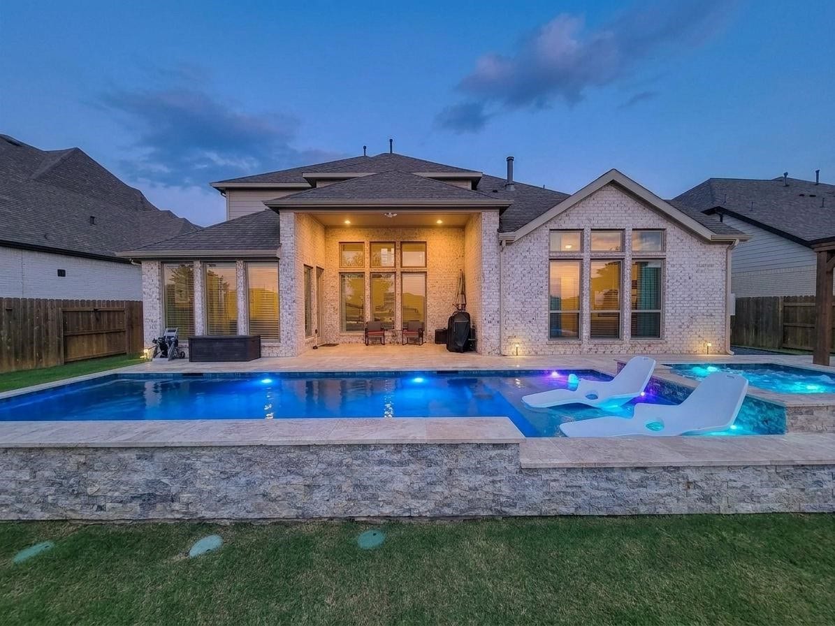Modern home with a backyard featuring a luxurious illuminated pool and spa. The exterior is elegantly designed with large windows and a covered patio area, ideal for outdoor entertaining.