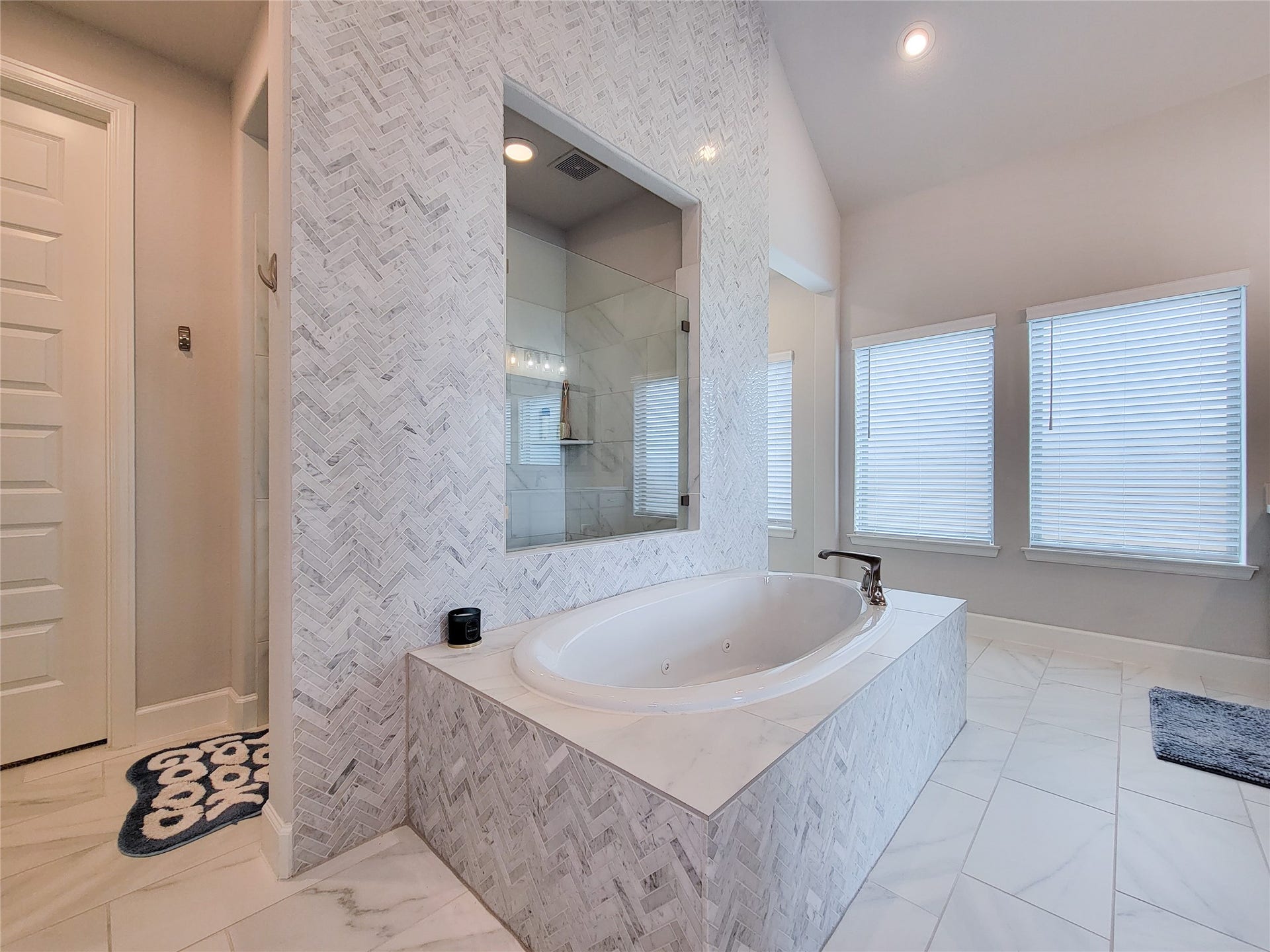 This bathroom features a luxurious, sunken tub with elegant marble tile surround and a glass-enclosed shower. The space is brightened by large windows with blinds, offering a modern and serene atmosphere.
