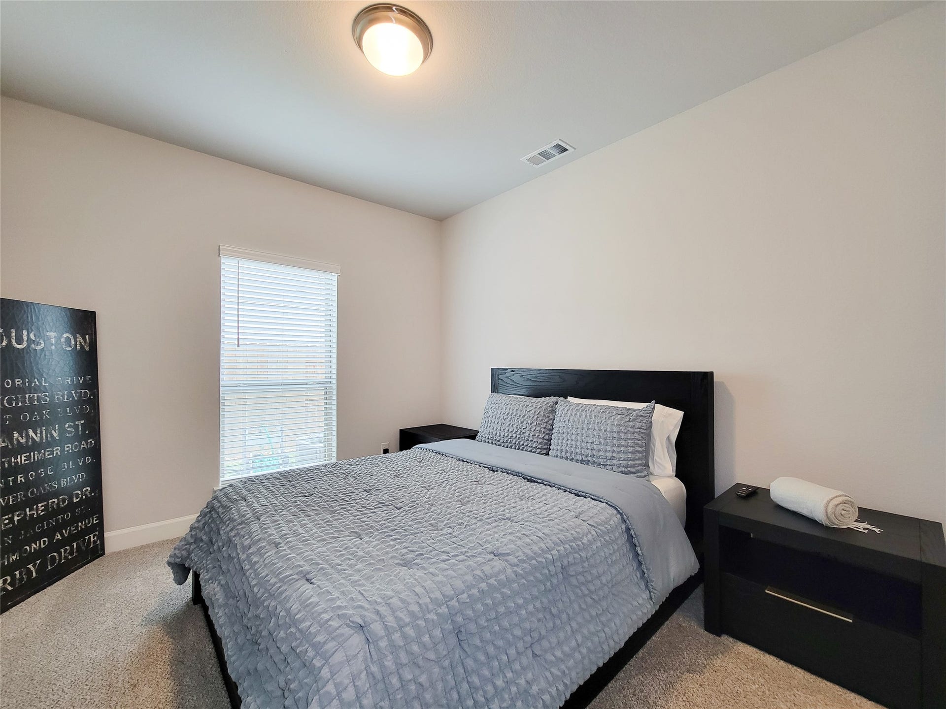 This bedroom features a cozy atmosphere with soft carpeting and neutral walls. A large window allows natural light to fill the space, complemented by a modern ceiling light.