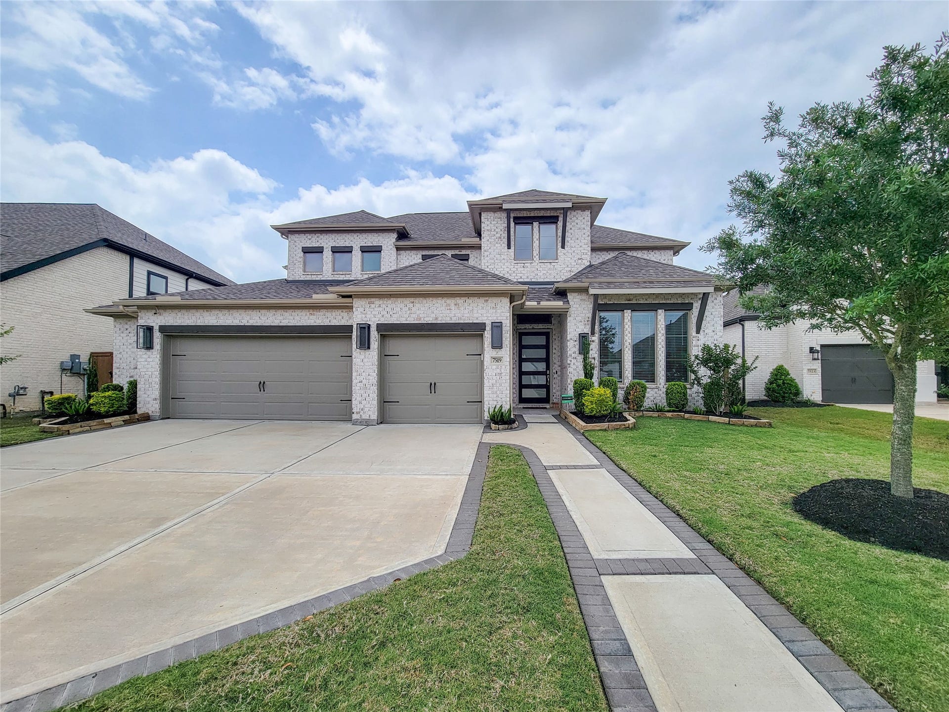This modern two-story home features a light brick exterior, a three-car garage, and a welcoming front path. The landscaped yard includes neatly trimmed shrubs and a lush lawn, enhancing the home's curb appeal.